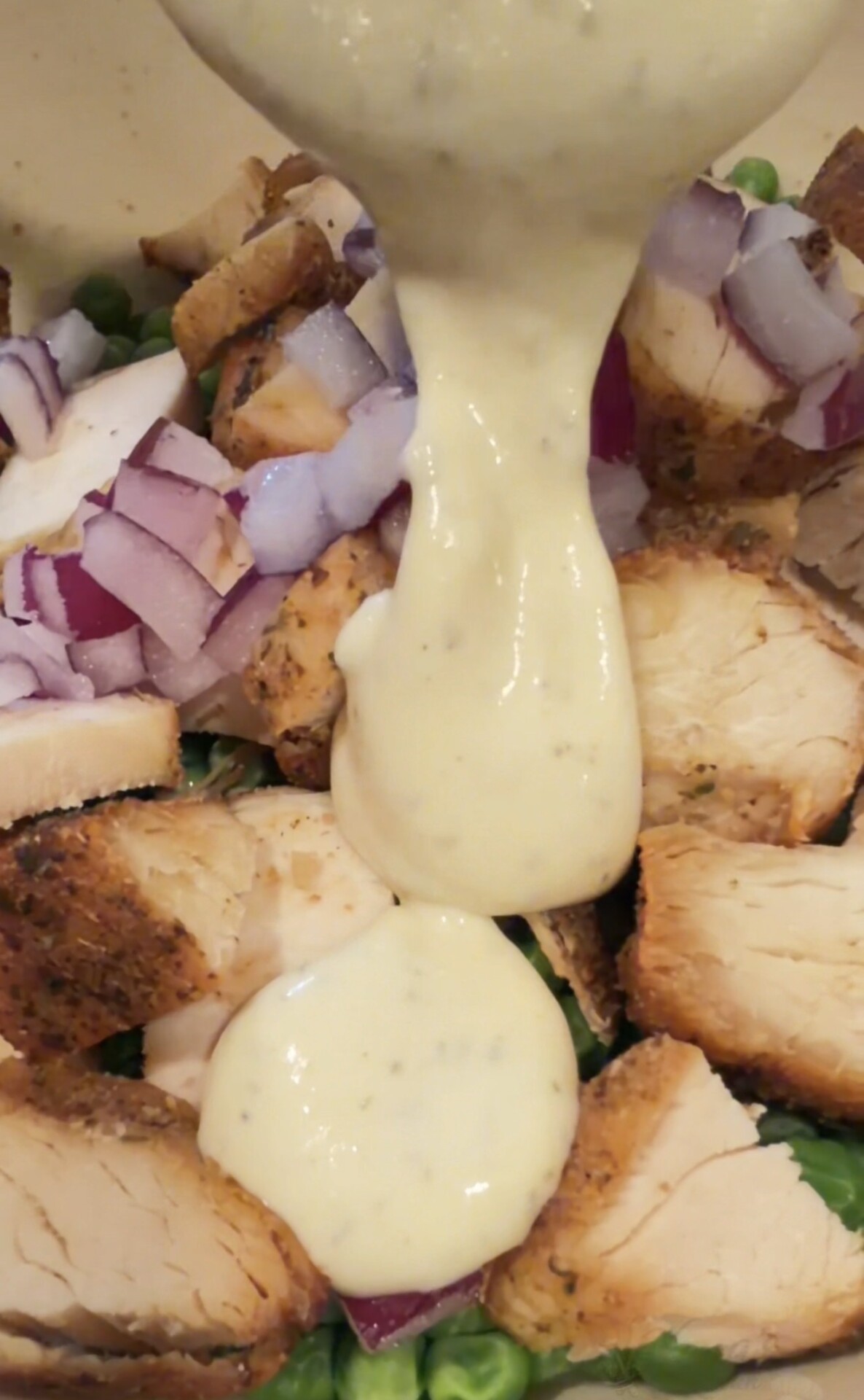 Creamy dressing being poured over a salad of sliced grilled chicken and chopped red onions.