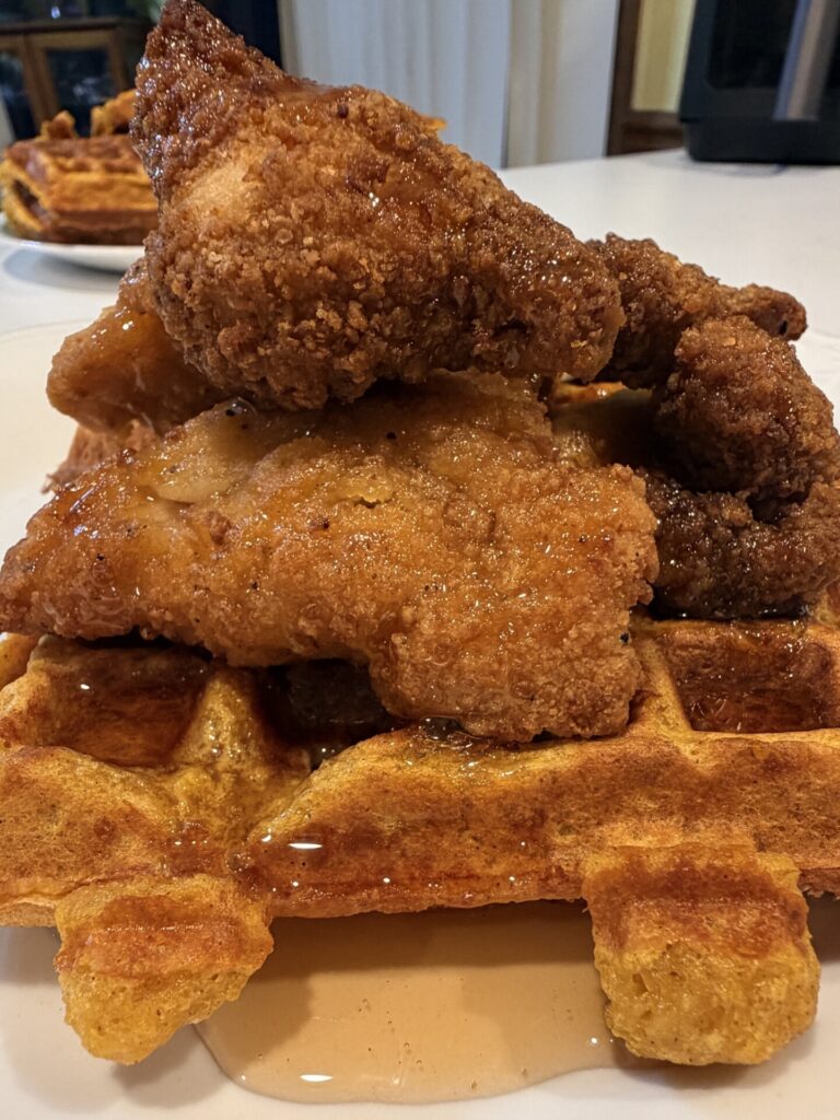 A large stack of crispy fried chicken pieces piled on top of a waffle, all drizzled with syrup.