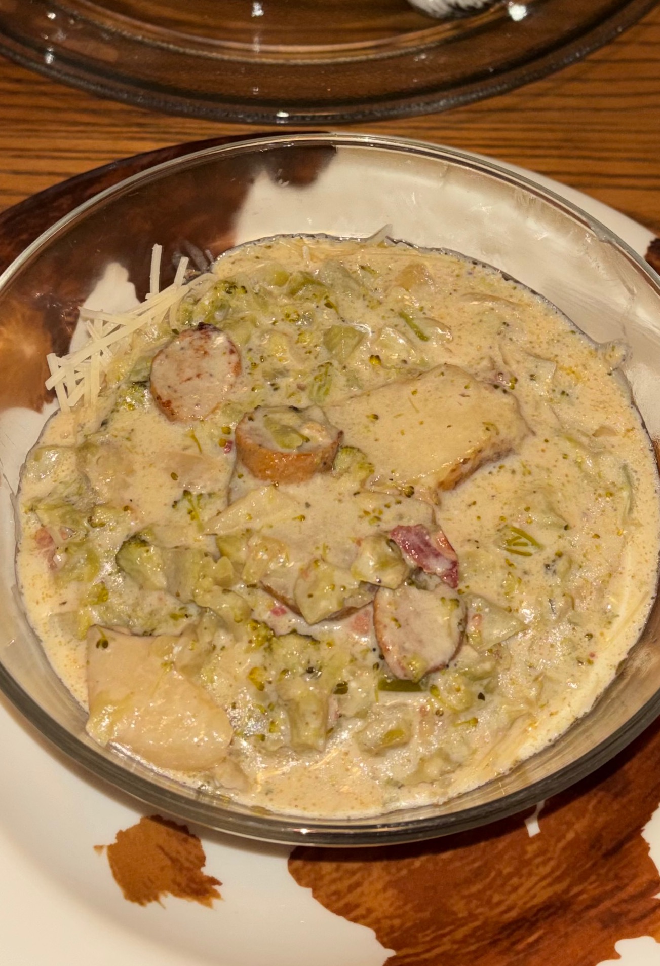 A glass bowl filled with creamy chicken and sausage stew, featuring pieces of meat and chopped vegetables, garnished with shredded cheese, placed on a plate.