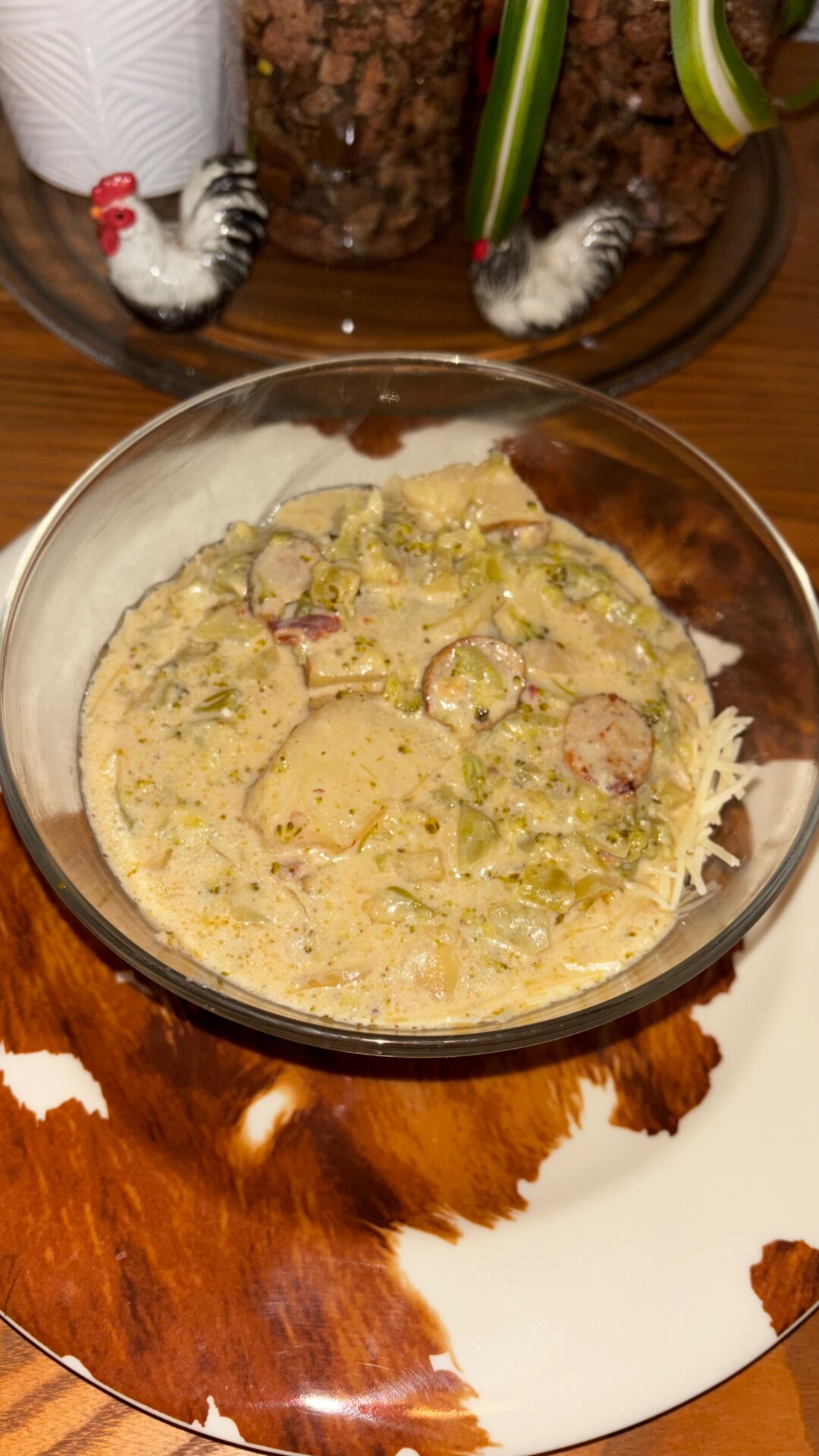 A bowl of creamy soup with sausage slices and vegetables sits on a brown-and-white plate, with rooster figurines and a plant in the background.