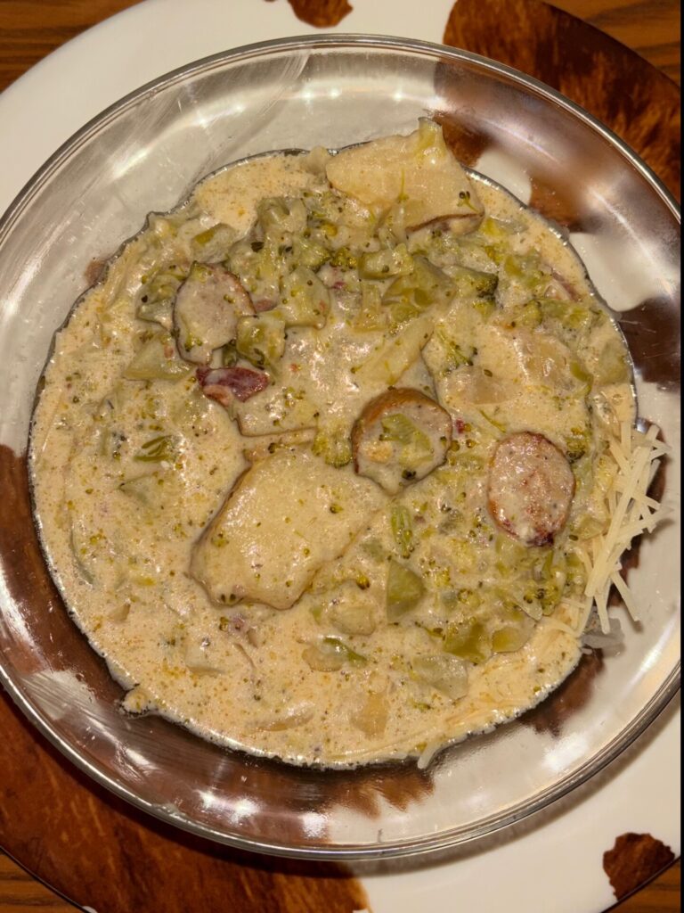 A bowl of creamy soup with potatoes, broccoli, sausage slices, and shredded cheese, served in a metal bowl on a brown and white plate.