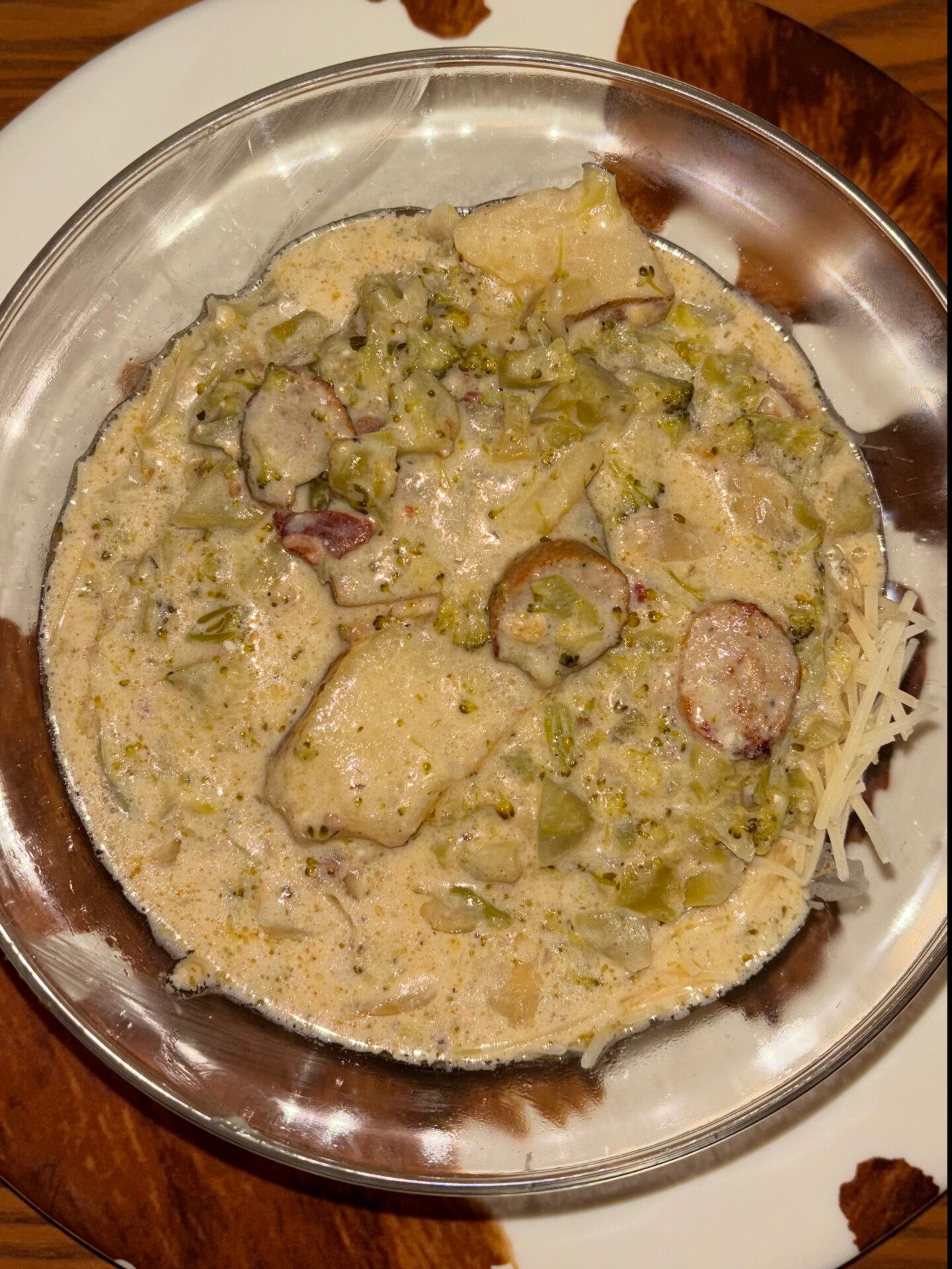 A bowl of creamy soup with potatoes, broccoli, sausage slices, and shredded cheese, served in a metal bowl on a brown and white plate.