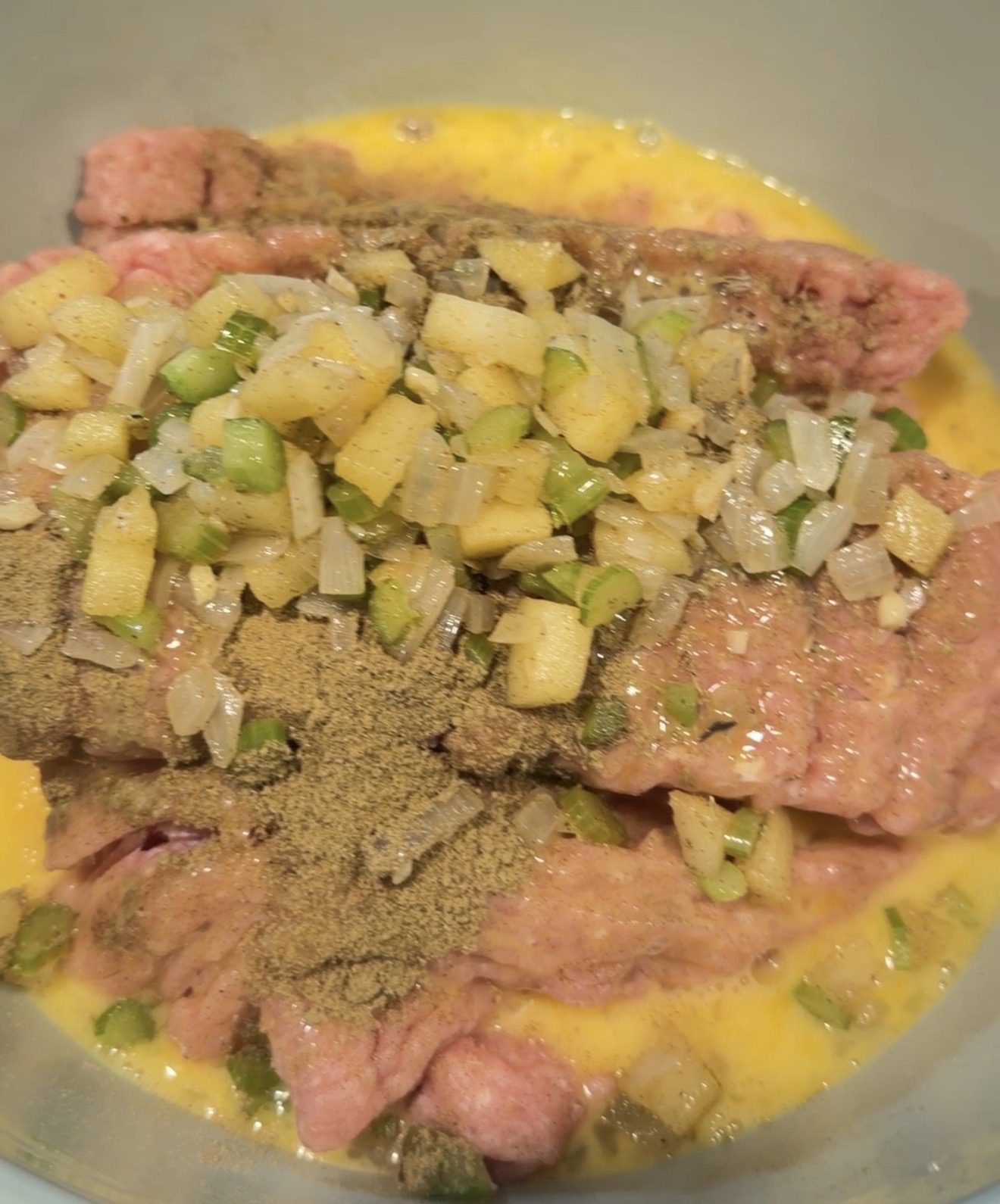 Raw ground meat in a bowl with beaten egg, chopped vegetables, and a mound of seasoning.