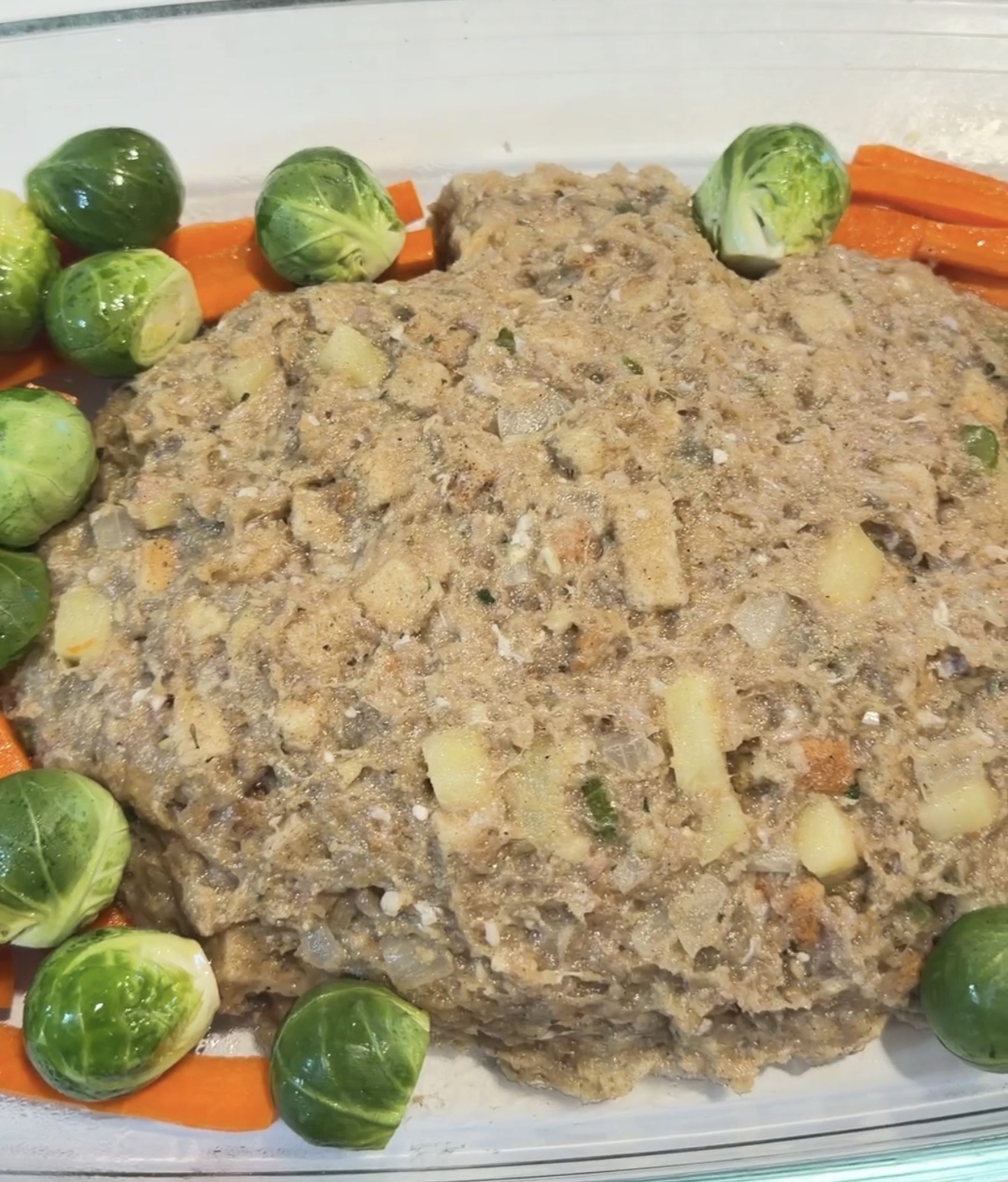 A glass baking dish containing a large portion of savory stuffing surrounded by Brussels sprouts and carrot sticks.