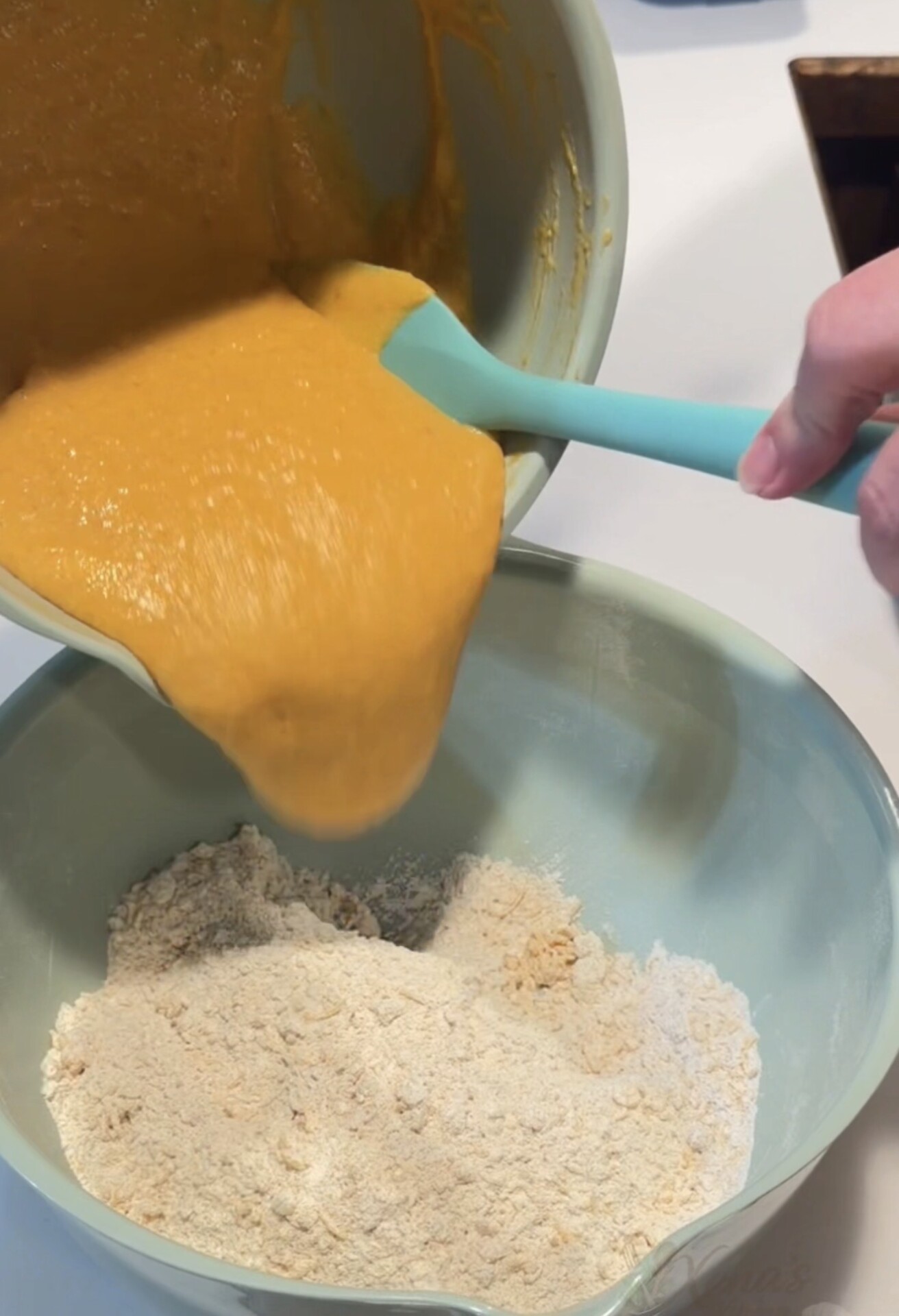 A person uses a spatula to pour orange batter from a mixing bowl into another bowl containing a dry flour mixture.