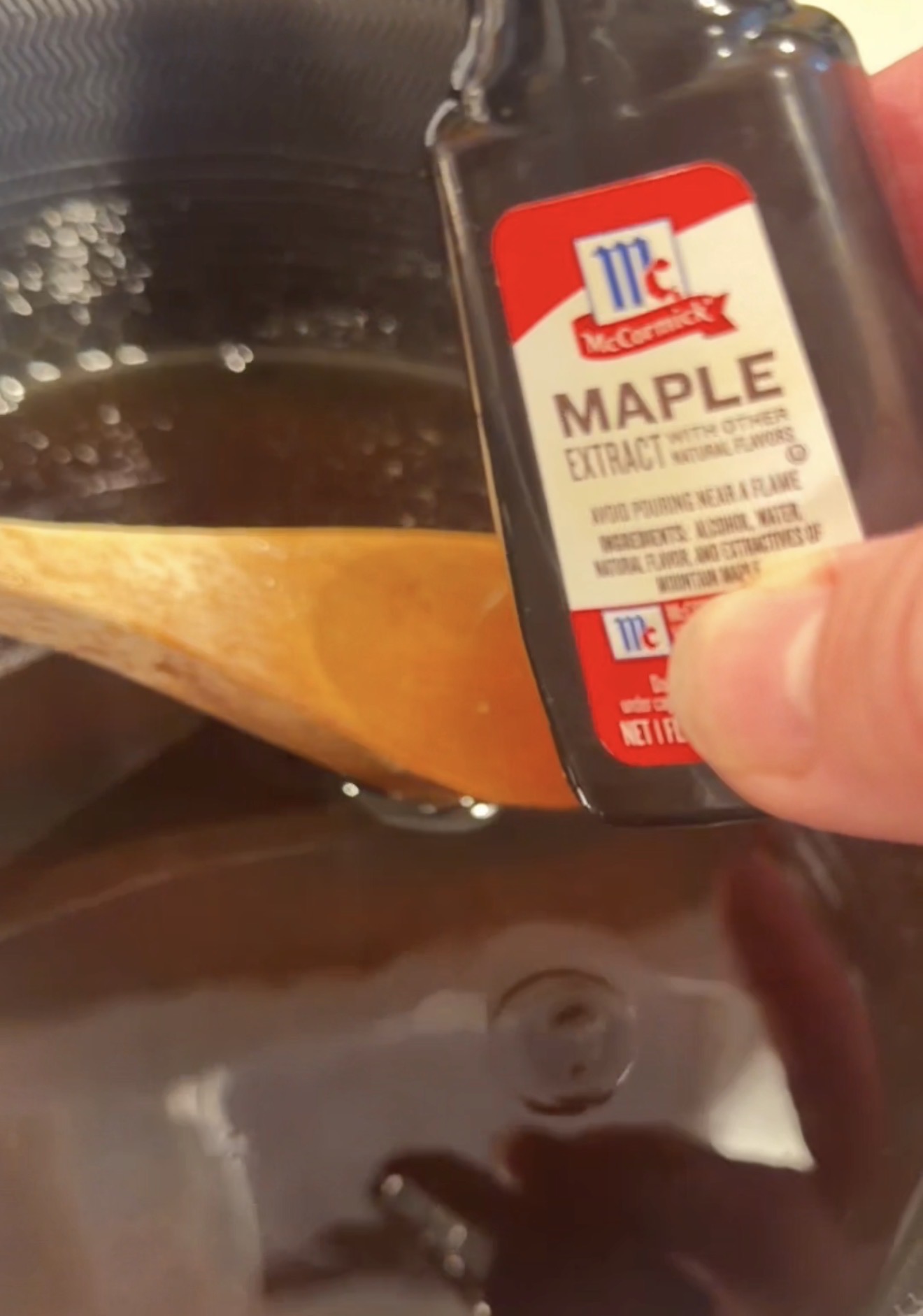 A hand pouring a bottle of McCormick maple extract into a dark liquid in a pot with a wooden spoon.