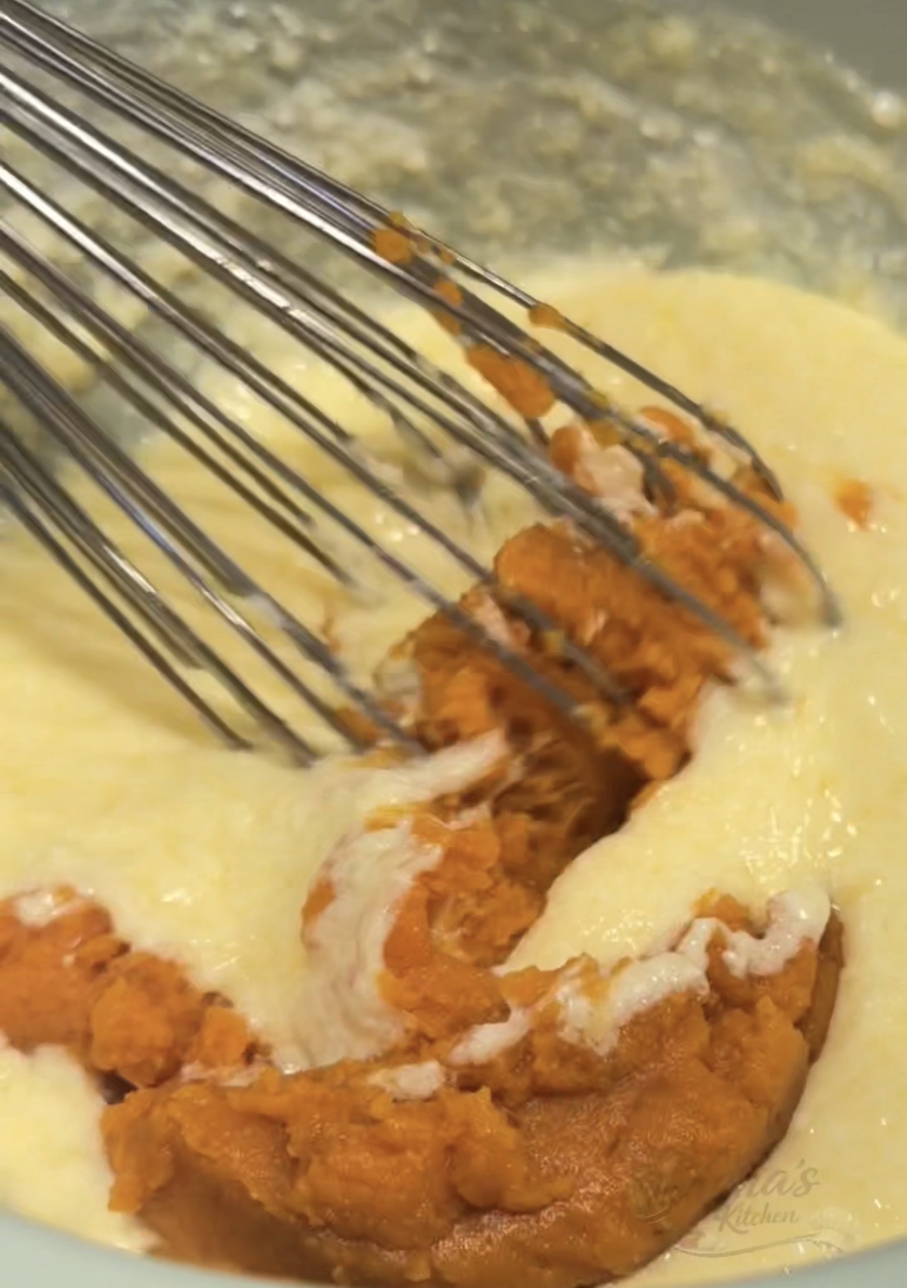 A metal whisk mixing orange pumpkin puree into a light-colored batter in a bowl.