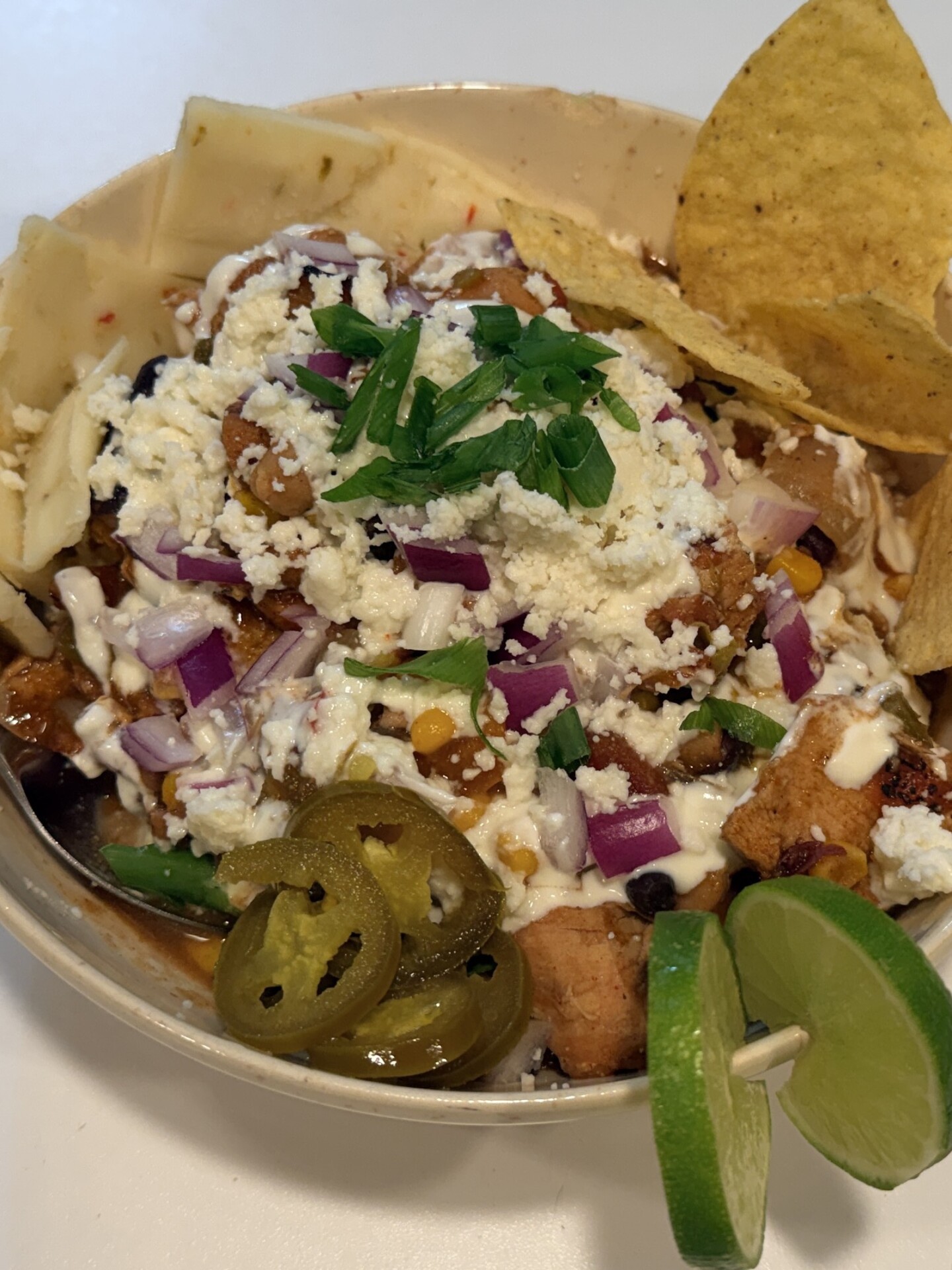 A bowl of chicken, beans, and corn topped with crumbled cheese, red onion, jalapeño slices, and green onions, served with tortilla chips and lime wedges.