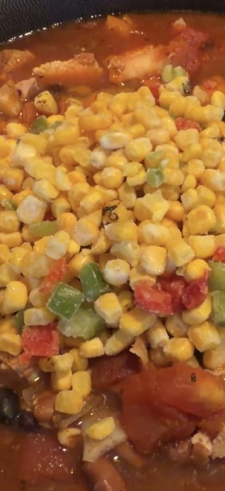 A close-up of a pot of chili topped with a large pile of frozen corn and diced bell peppers.