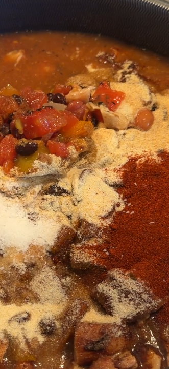 A pot of cooking stew topped with heaps of diced tomatoes, black beans, red chili powder, and onion powder.