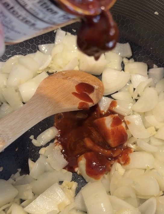 Red sauce being poured from a container onto a wooden spoon and sautéing onions in a pan.