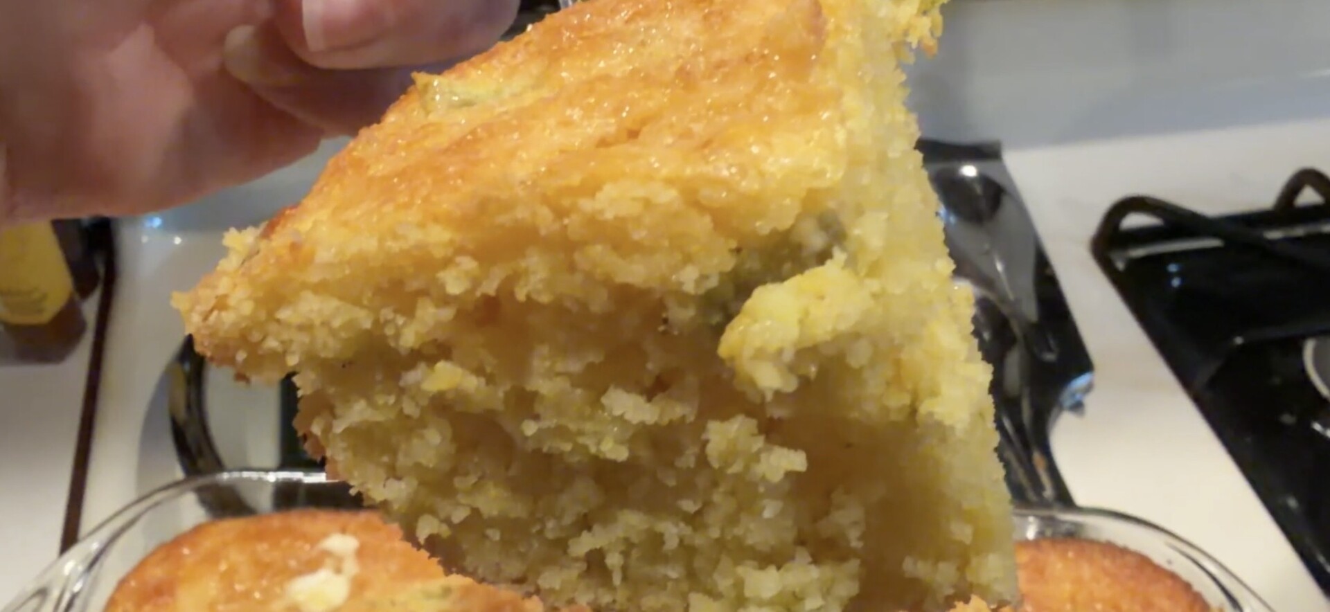 A hand holds a golden, crumbly wedge of cornbread above a baking dish.