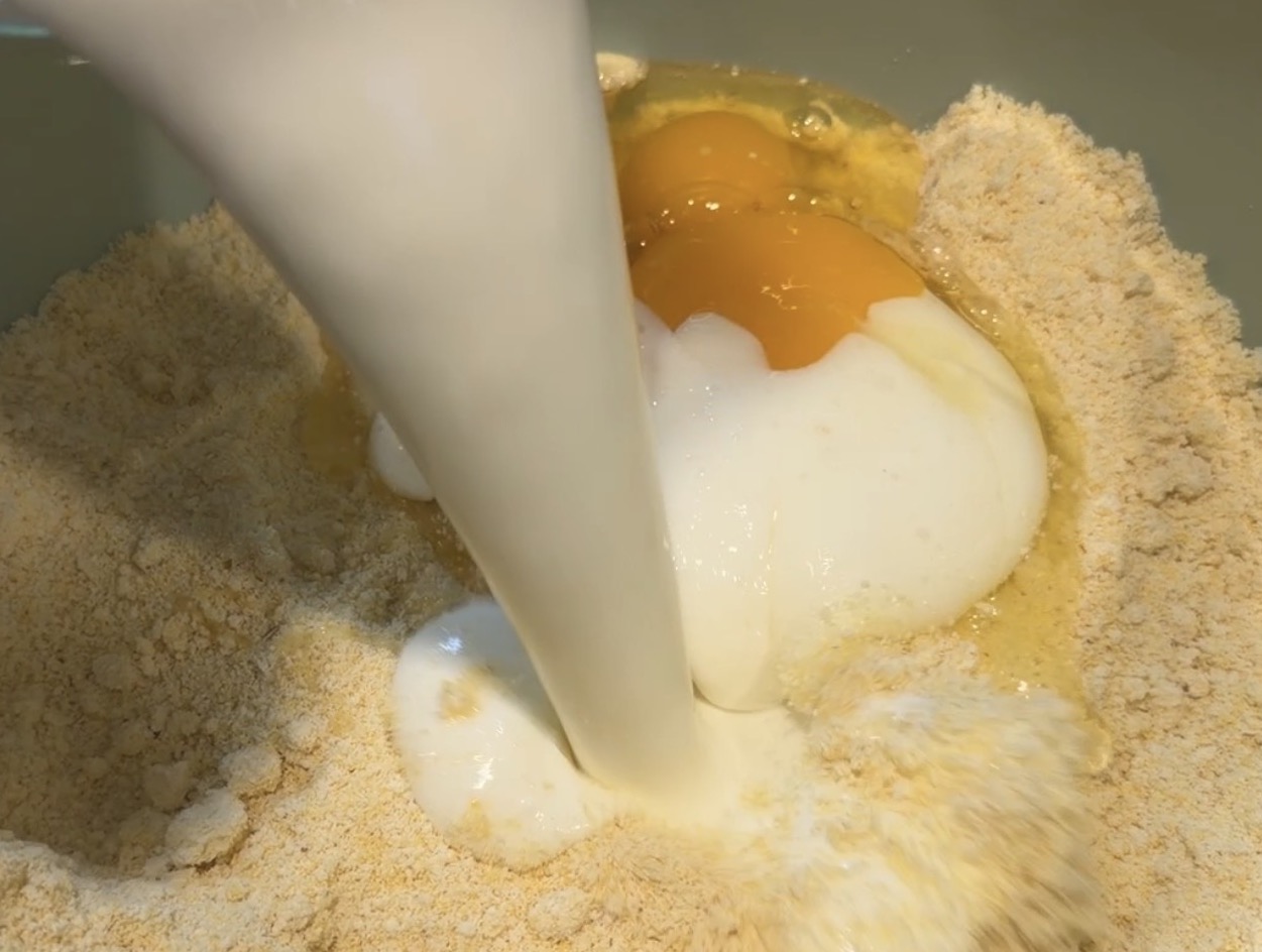 Milk being poured into a bowl containing flour and cracked raw eggs.
