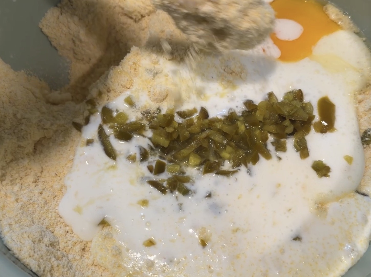 A mixing bowl containing cornmeal, a cracked egg, milk, and chopped green jalapeños.