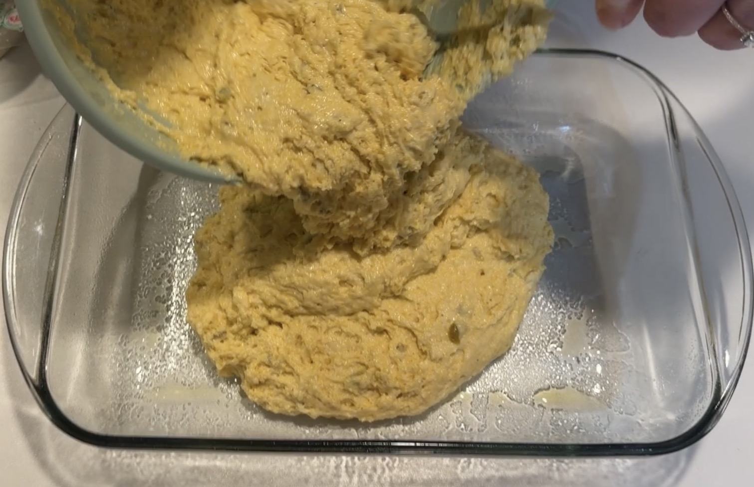 Thick yellow batter being poured from a bowl into a greased glass baking dish.