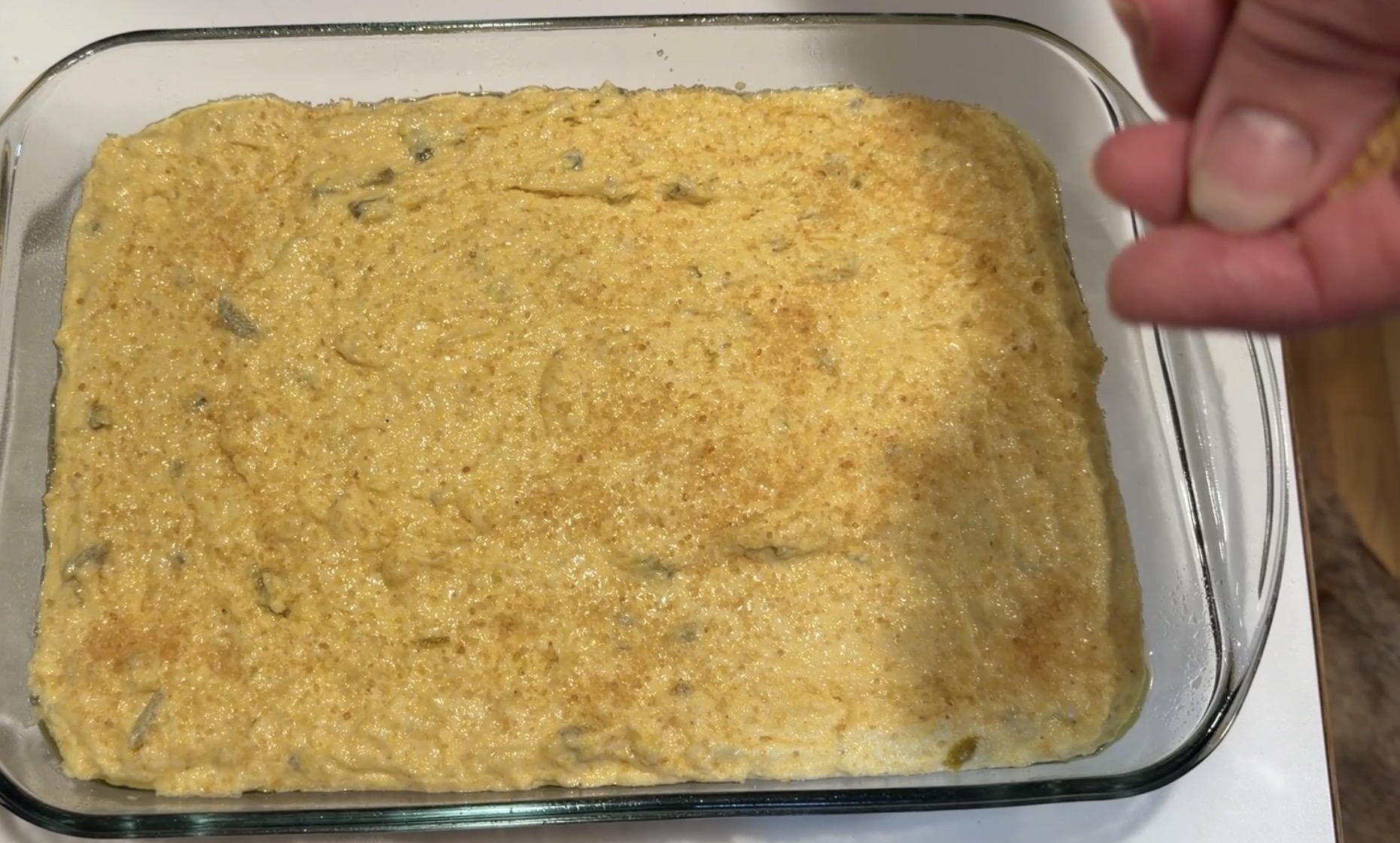 A hand sprinkles seasoning over a yellow corn casserole in a glass baking dish.