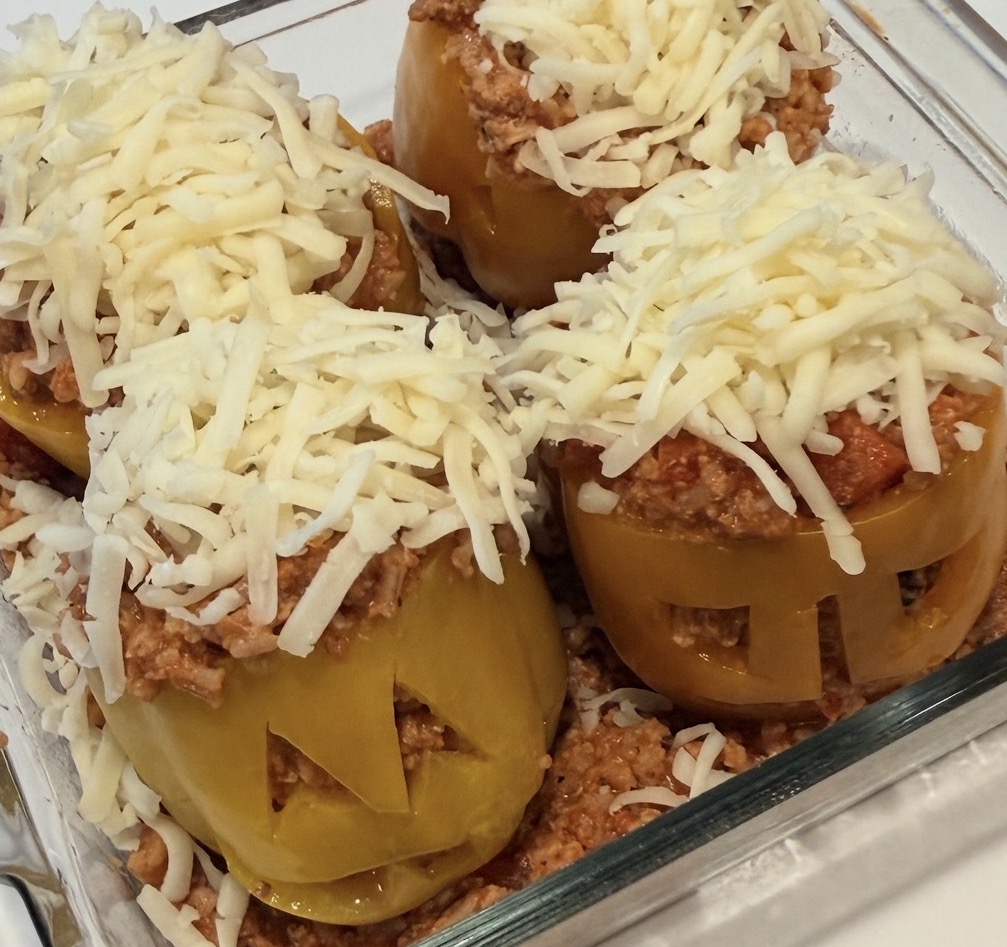 Four yellow bell peppers carved like jack-o-lanterns, filled with meat sauce and topped with shredded cheese in a glass baking dish.