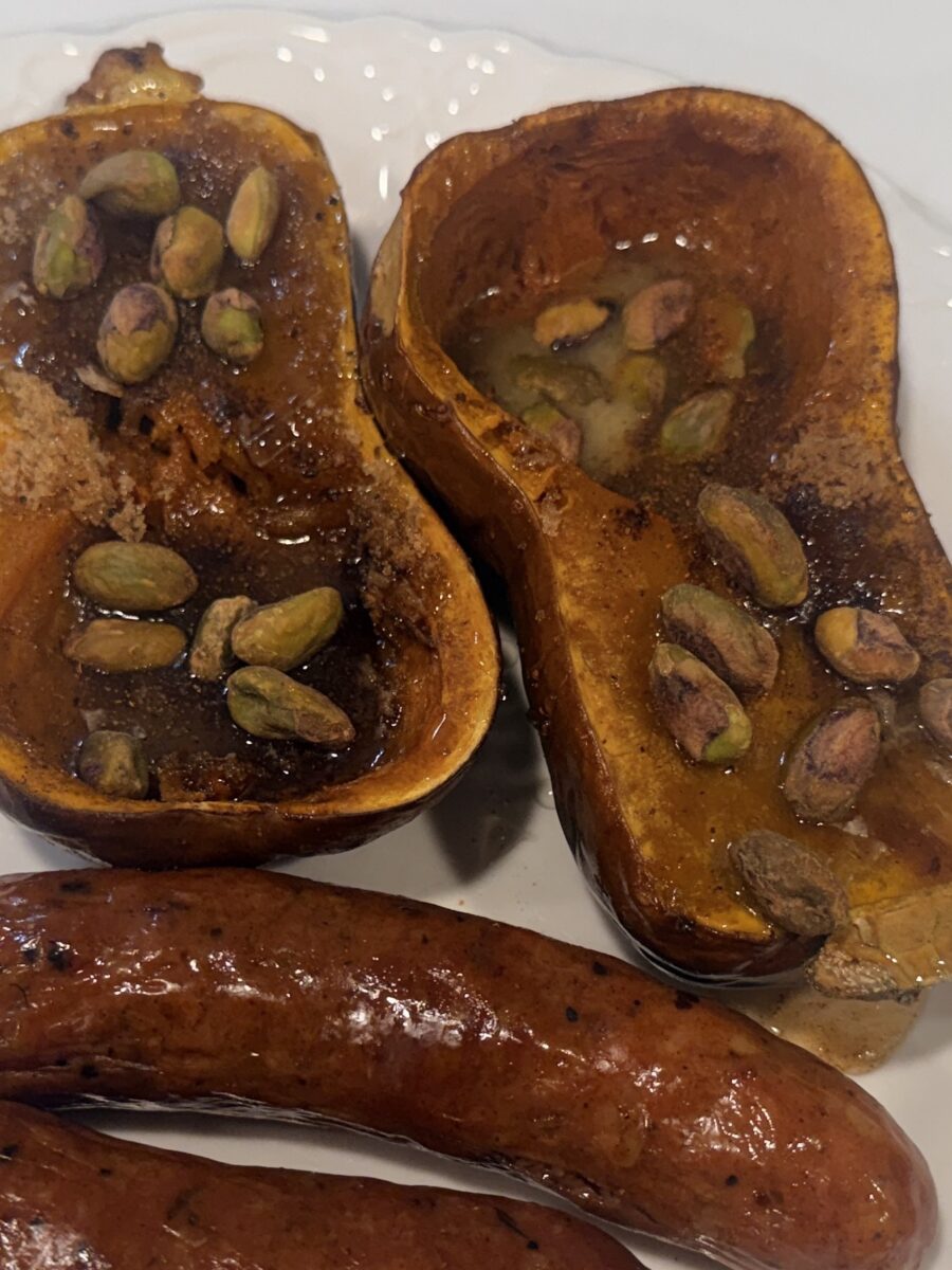 Two halves of roasted butternut squash topped with pistachios and brown sugar, served with sausages on a plate.