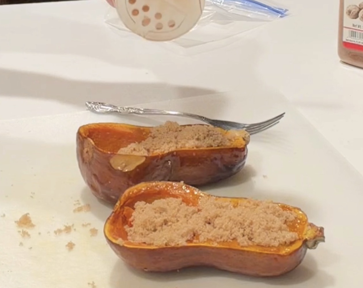 A hand shakes brown sugar from a spice jar onto two halves of roasted butternut squash.