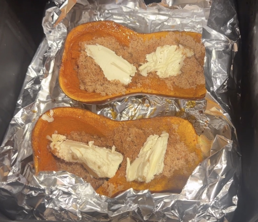 Two halves of a butternut squash, roasted and topped with brown sugar and pats of butter, sitting on aluminum foil in an air fryer basket.
