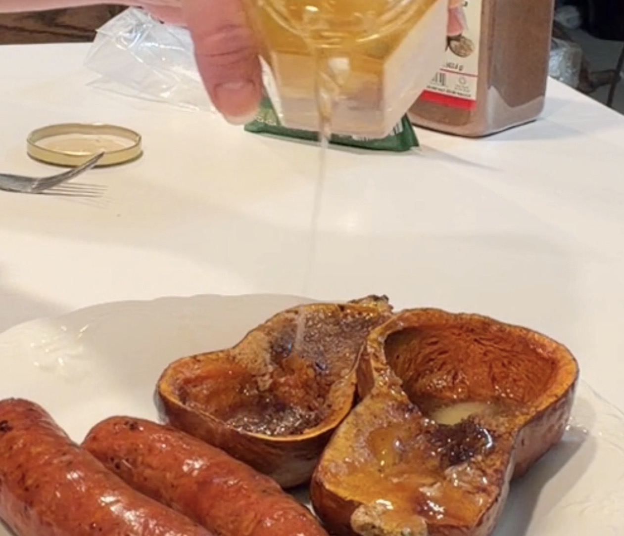 Honey being drizzled onto two halves of roasted butternut squash next to two sausages on a white plate.