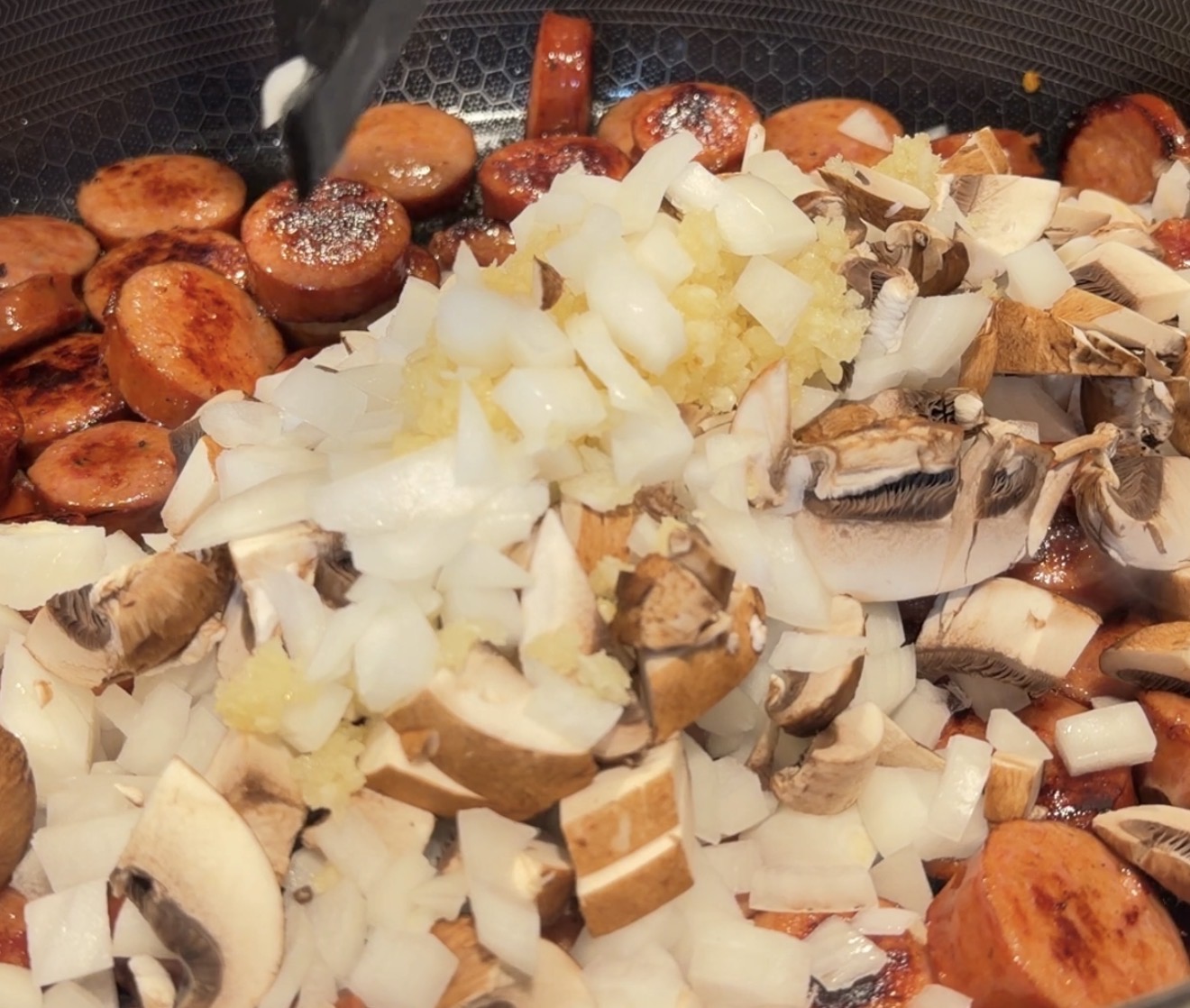 Sliced sausage, chopped onions, minced garlic, and sliced mushrooms cooking in a pan.