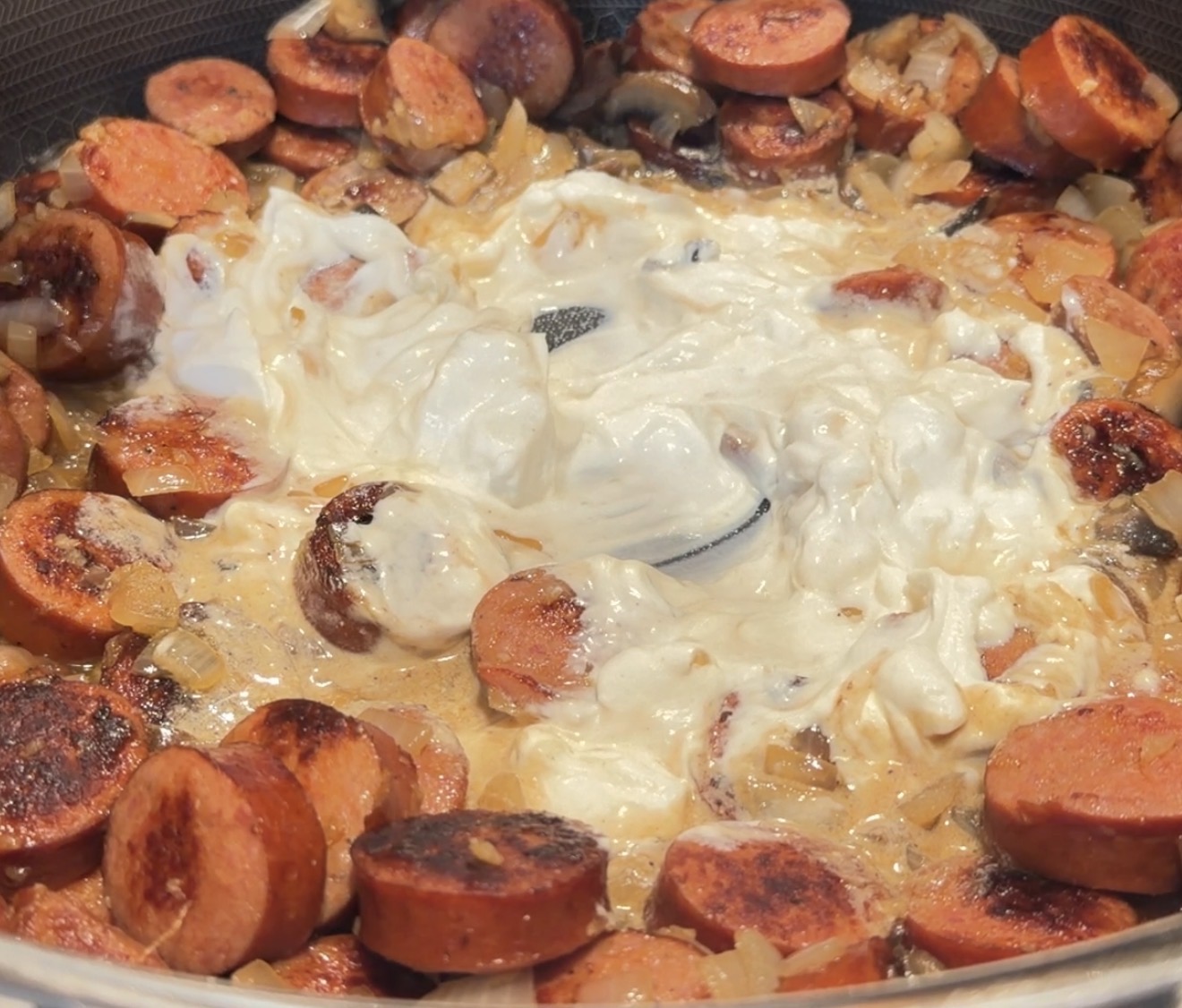 Sautéed sliced sausage and onions in a creamy white sauce cooking in a pan.