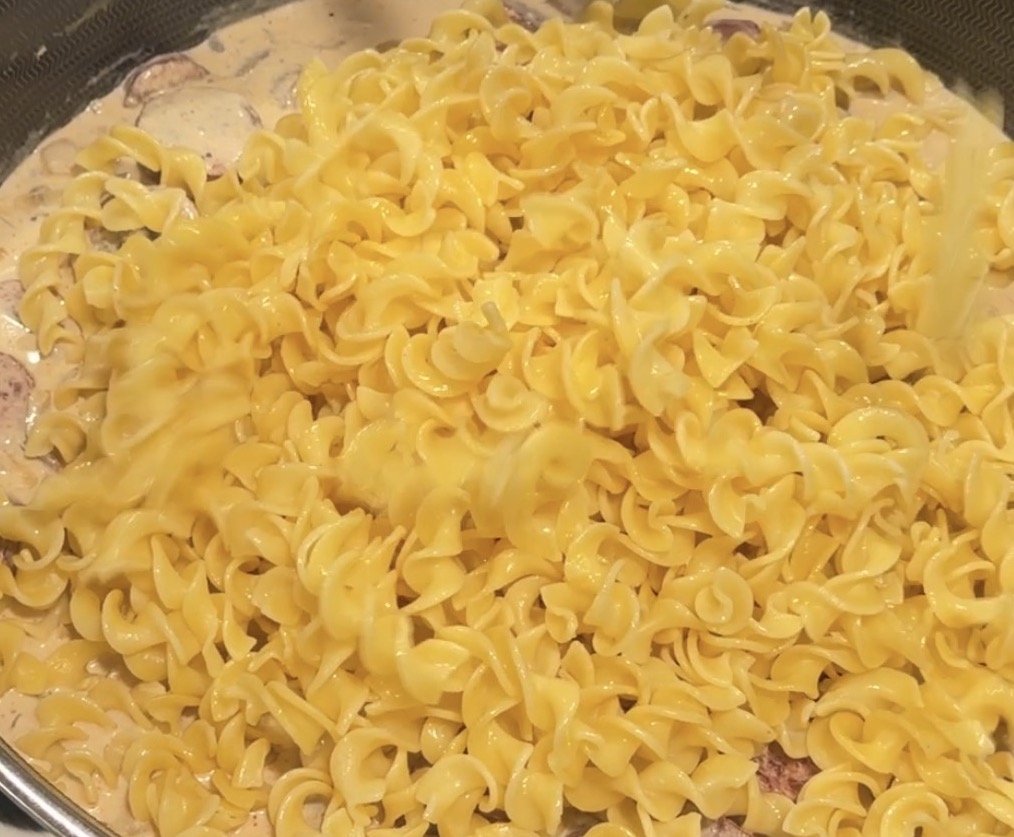 Spiral egg noodles being stirred into a creamy sauce with meat in a pan.