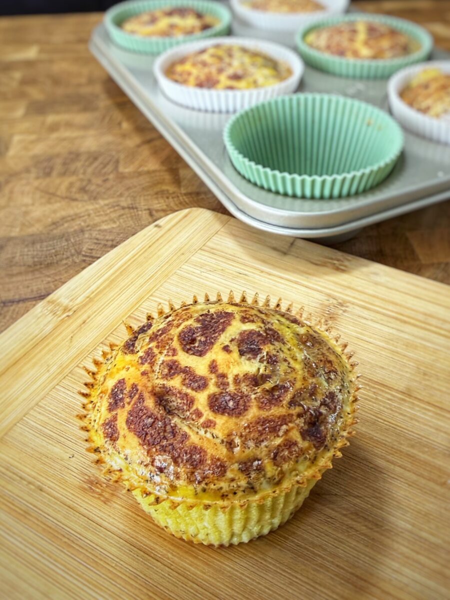 A baked egg muffin in a paper liner sits on a wooden board, with a muffin tin holding more egg muffins and empty green liners in the background.