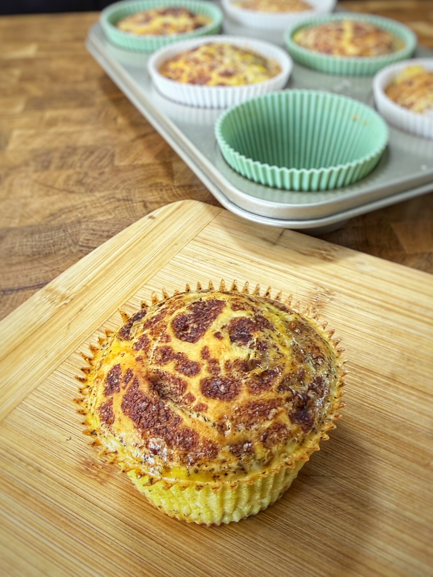 A baked egg muffin in a paper liner sits on a wooden board, with a muffin tin holding more egg muffins and empty green liners in the background.