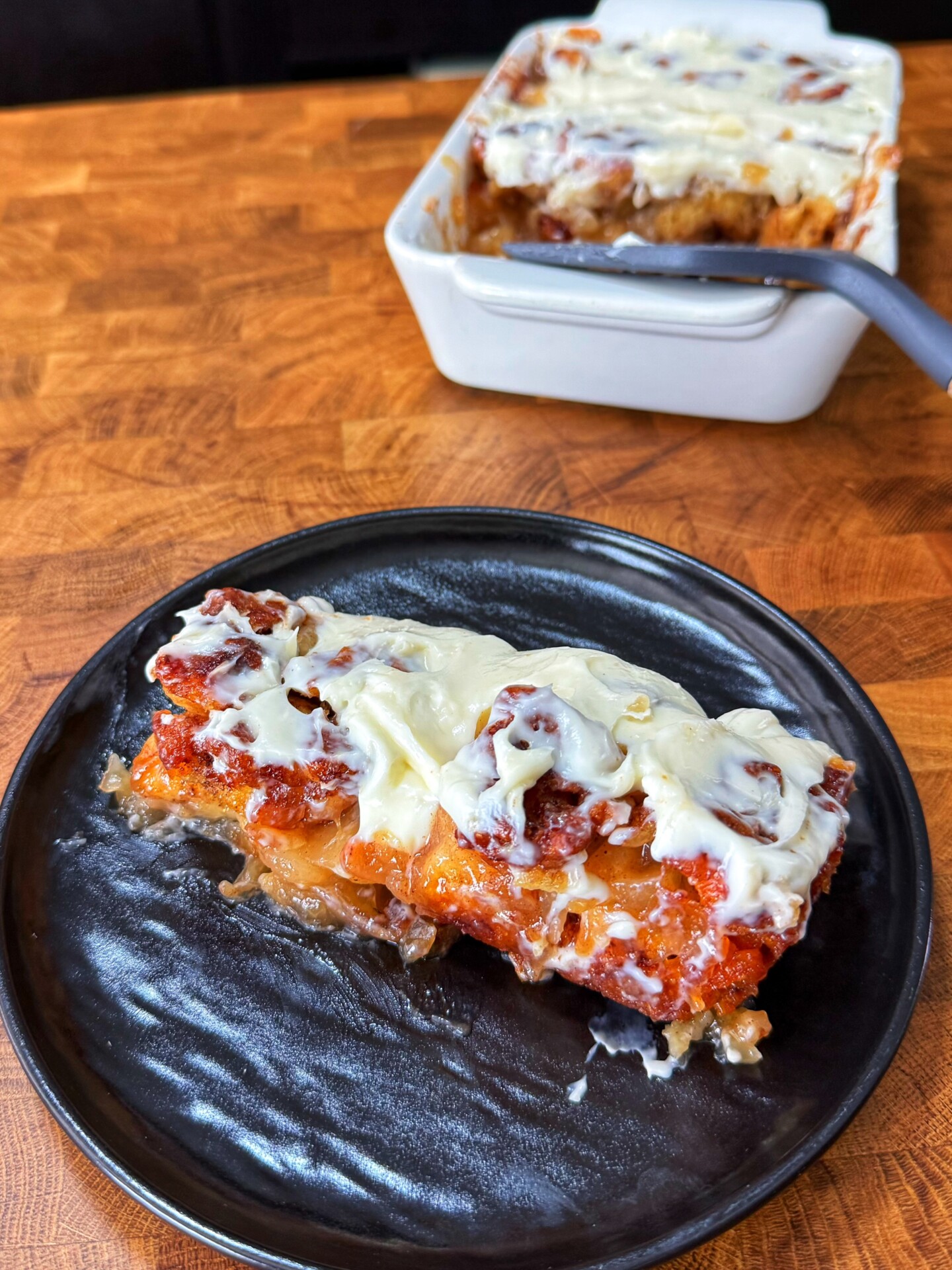 A slice of cheesy baked dish on a black plate, with the rest of the dish in a white baking pan in the background, both sitting on a wooden surface.