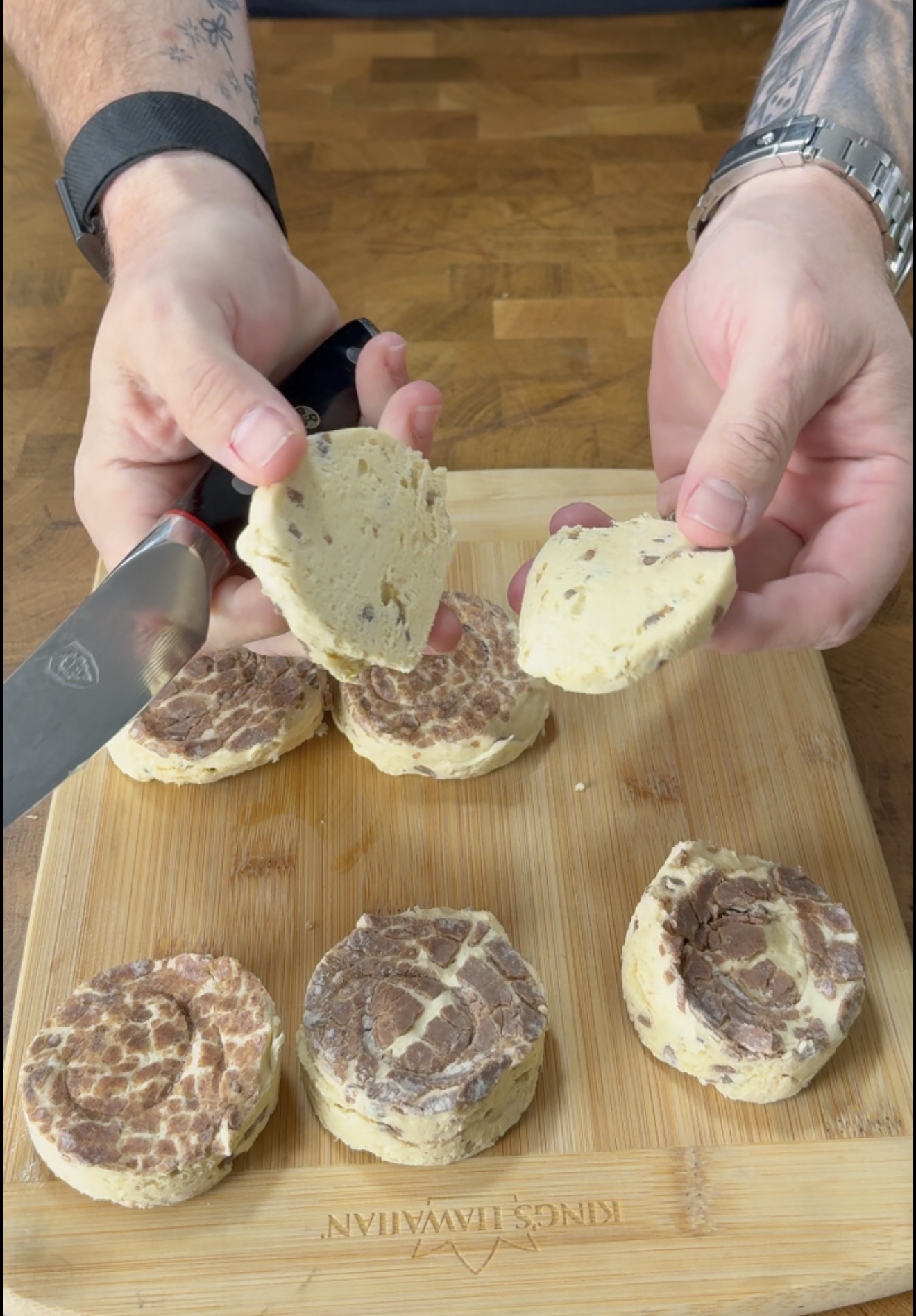 A person slices marbled cookie dough on a wooden cutting board, holding one round slice in each hand. Several dough slices and a knife are also visible on the board.