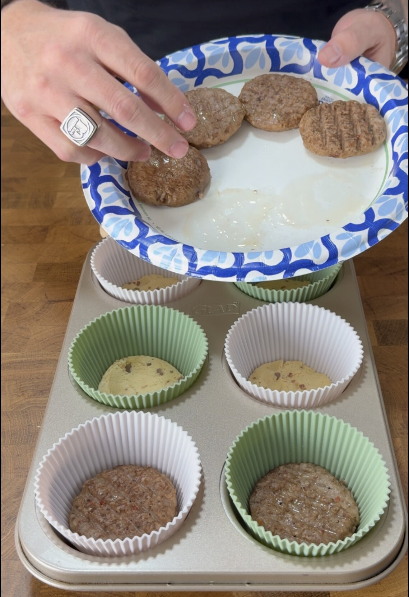 A person places cooked sausage patties into muffin cups filled with batter in a muffin tin. The sausage patties are on a blue and white paper plate.