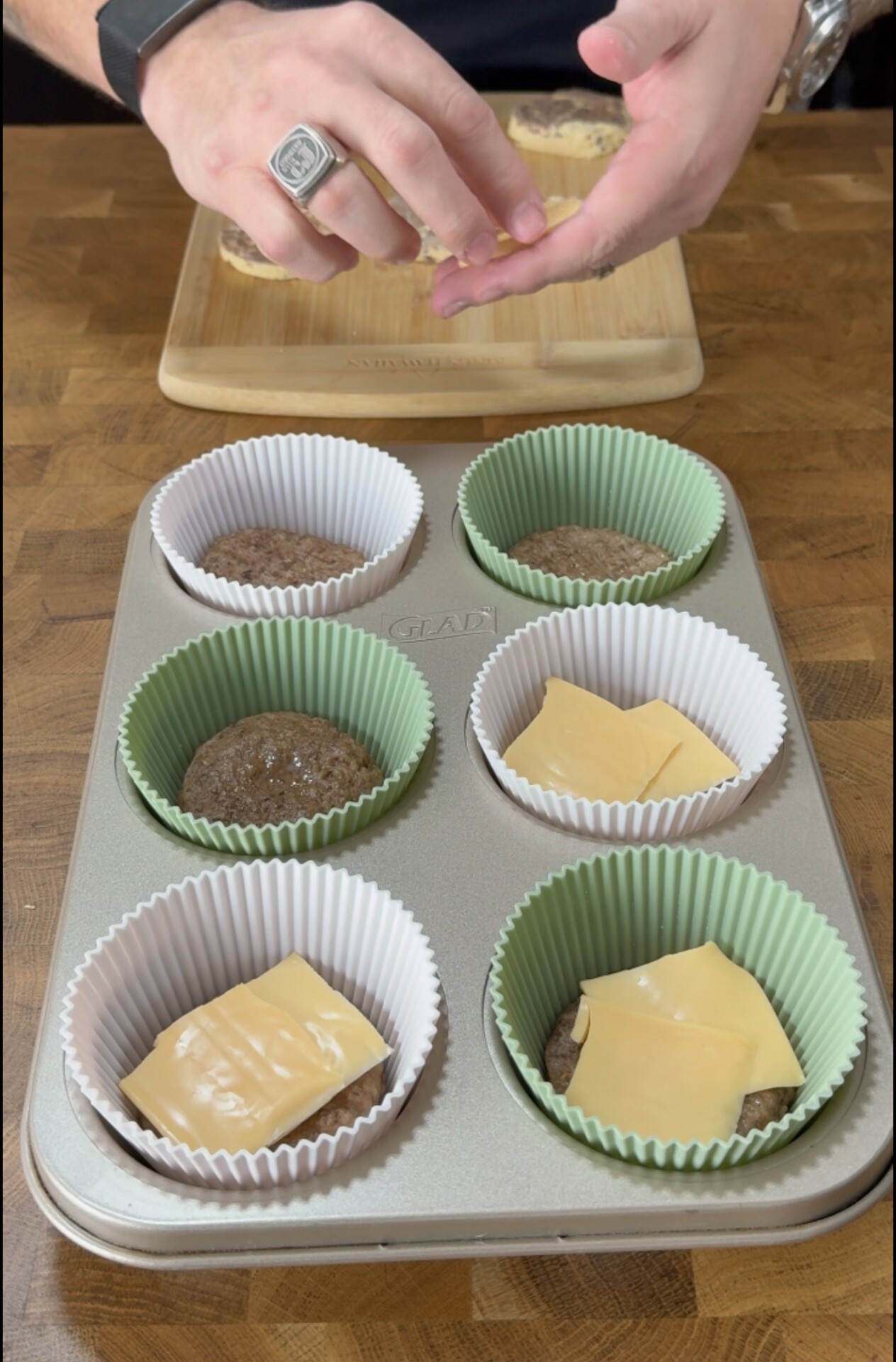 A person prepares muffin tin sliders, placing cheese slices on top of cooked patties inside a six-cup muffin pan lined with green and white paper liners. A wooden board with bread is in the background.