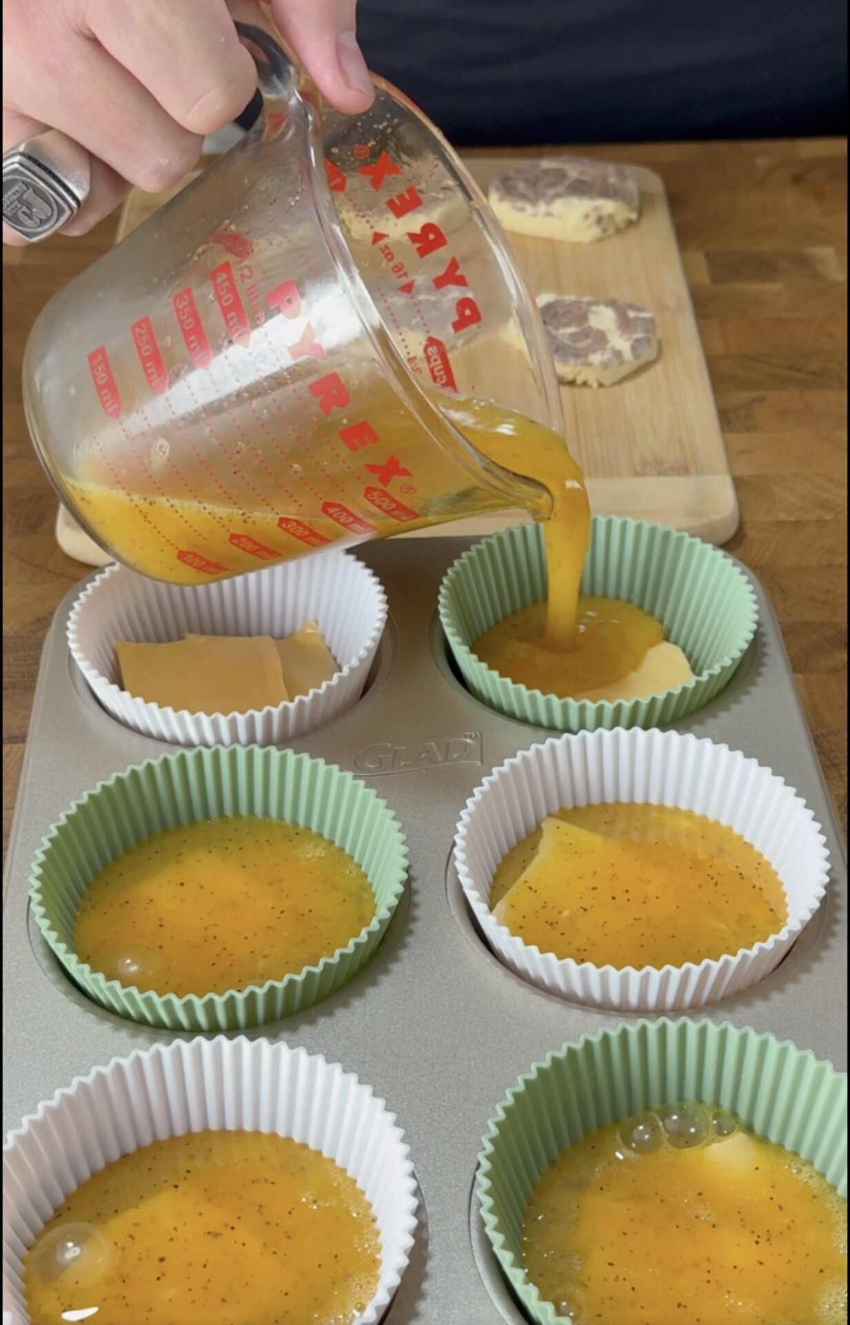 A hand pours orange egg mixture from a glass measuring cup into green and white muffin liners in a muffin tin. Slices of dough or pastry are on a wooden board in the background.