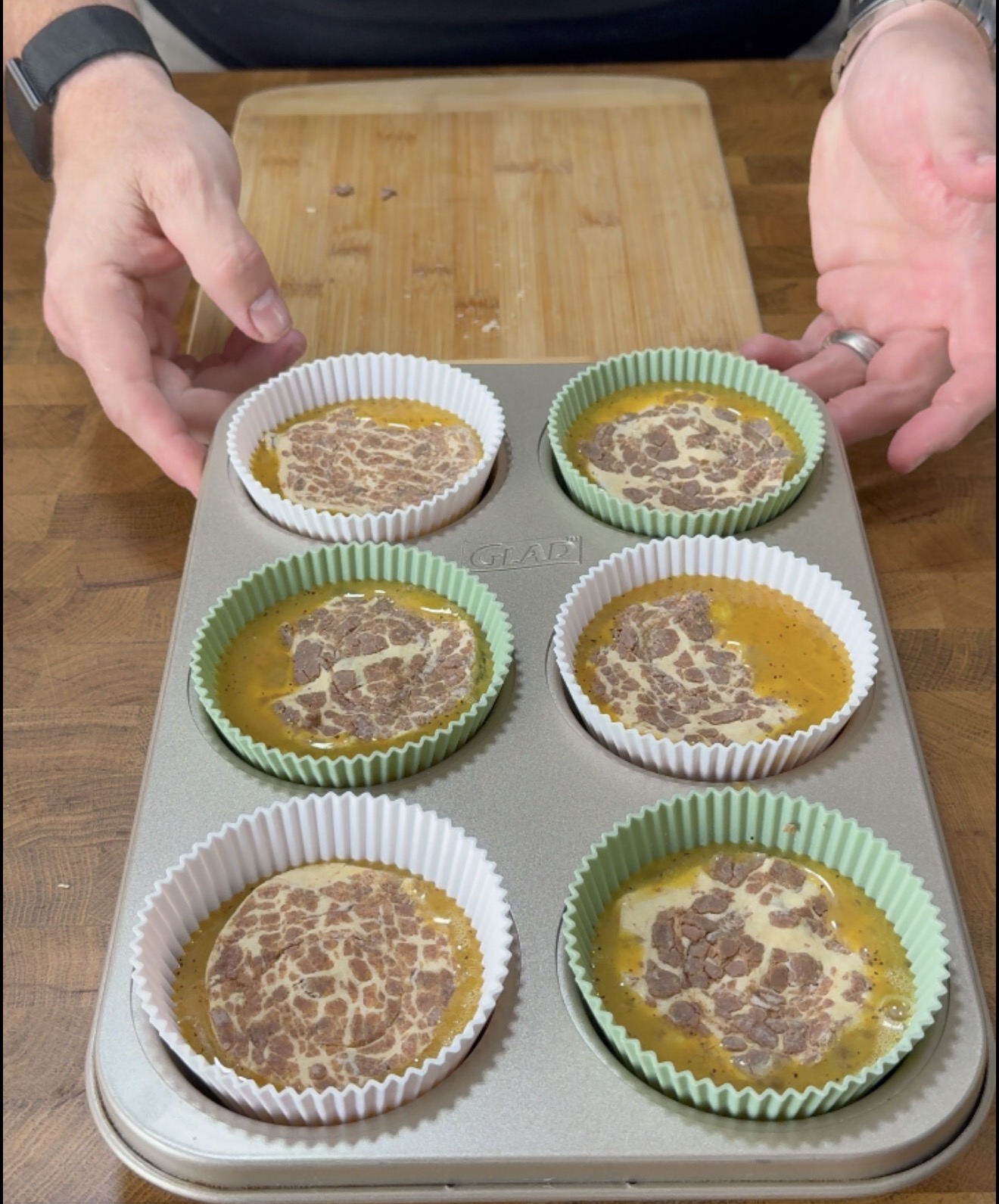 A person prepares six muffin liners in a metal tray, each filled with whisked eggs and a slice of patterned sausage on top, set on a wooden cutting board.