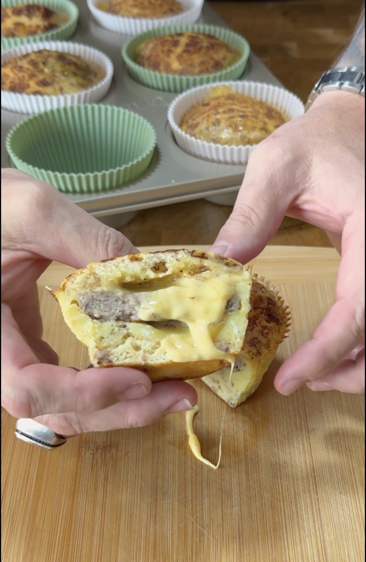 Close-up of hands pulling apart a muffin-shaped egg bite with sausage and melted cheese inside. More egg bites in a muffin pan are visible in the background.