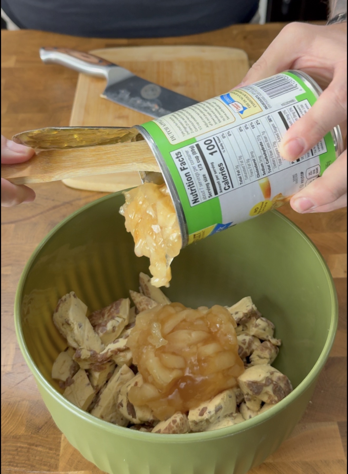 A person pours canned apple pie filling into a green bowl filled with chopped chocolate chip cookies. A cutting board and knife are visible in the background.