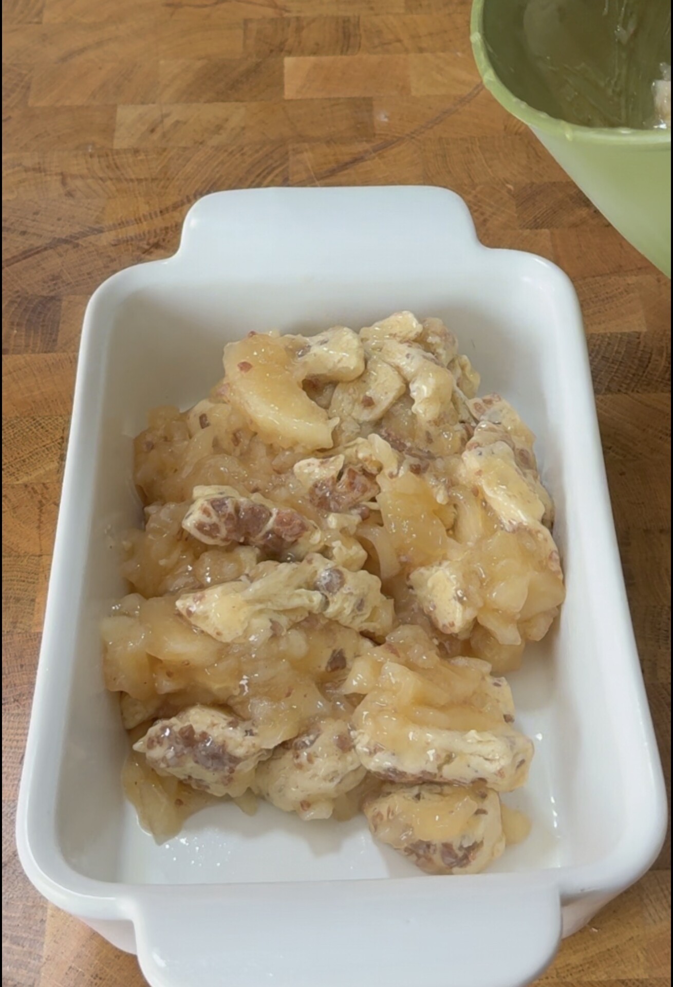 A white baking dish filled with a mixture of chopped apples and nuts in a syrupy sauce sits on a wooden countertop. A green bowl with remnants of batter is partially visible nearby.