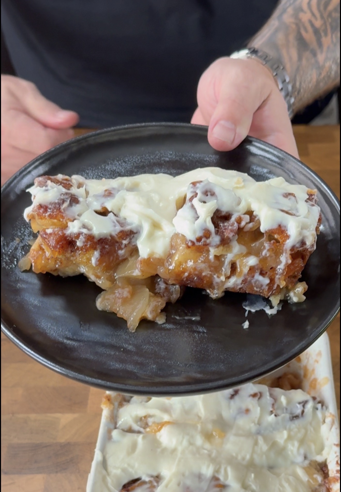 A person holds a black plate with a piece of frosted bread pudding, showing a gooey, cinnamon-filled interior and creamy white icing on top.