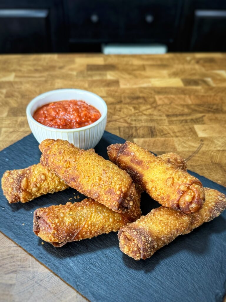 A pile of fried egg rolls on a slate board next to a small bowl of red dipping sauce.