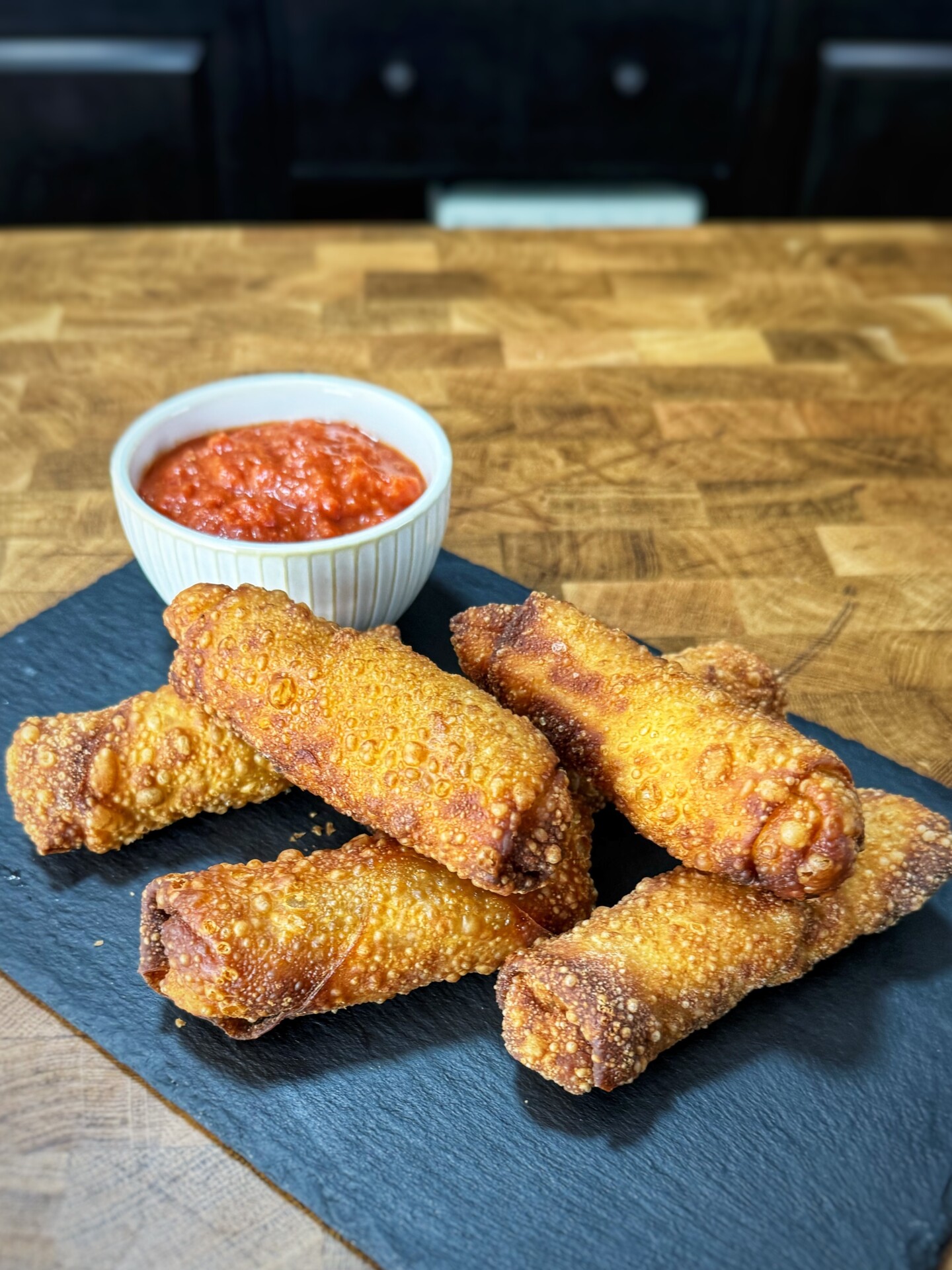 A pile of fried egg rolls on a slate board next to a small bowl of red dipping sauce.