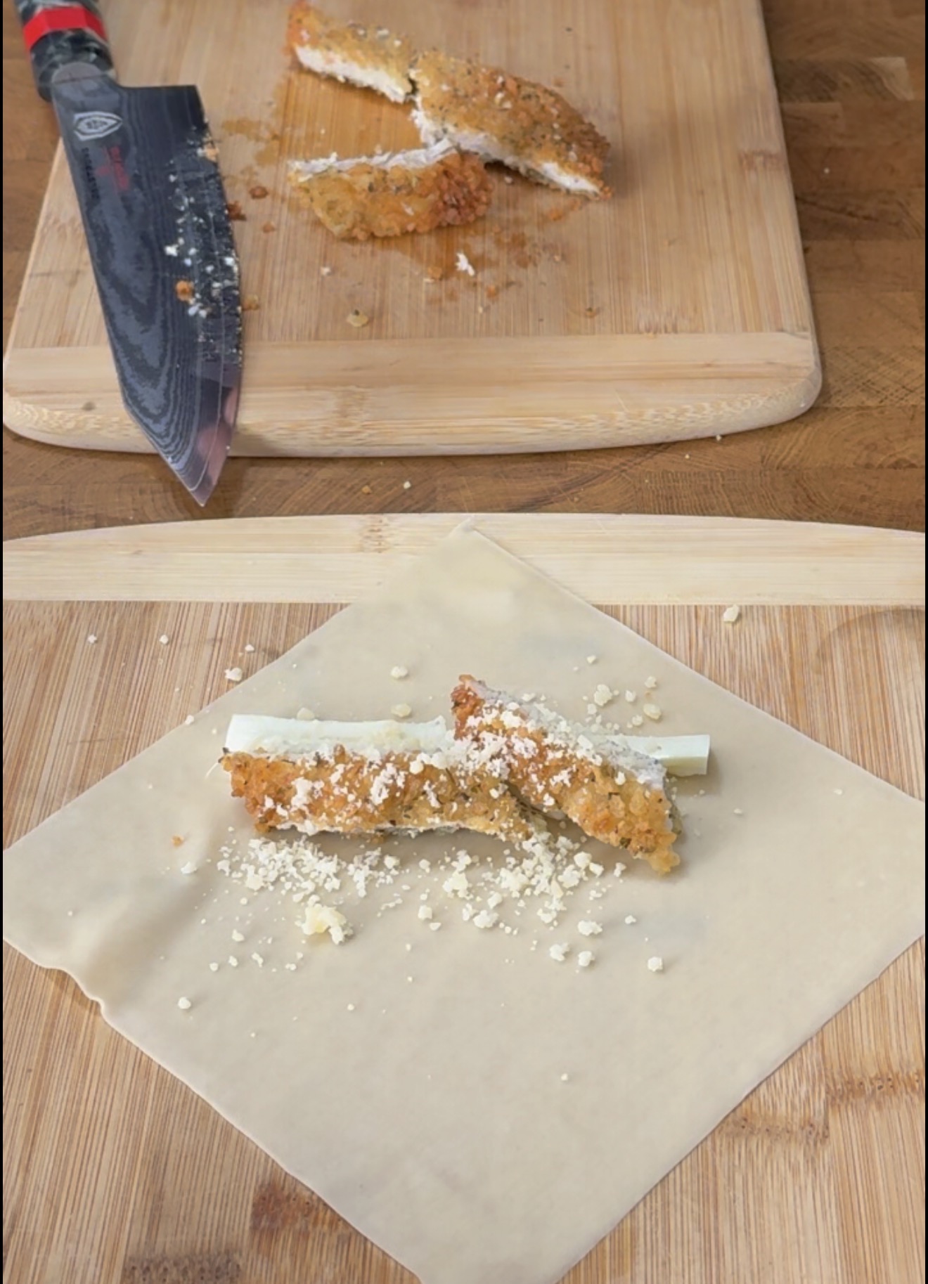 A piece of breaded chicken and a stick of cheese placed on a square egg roll wrapper, with a knife and more chicken on a cutting board nearby.