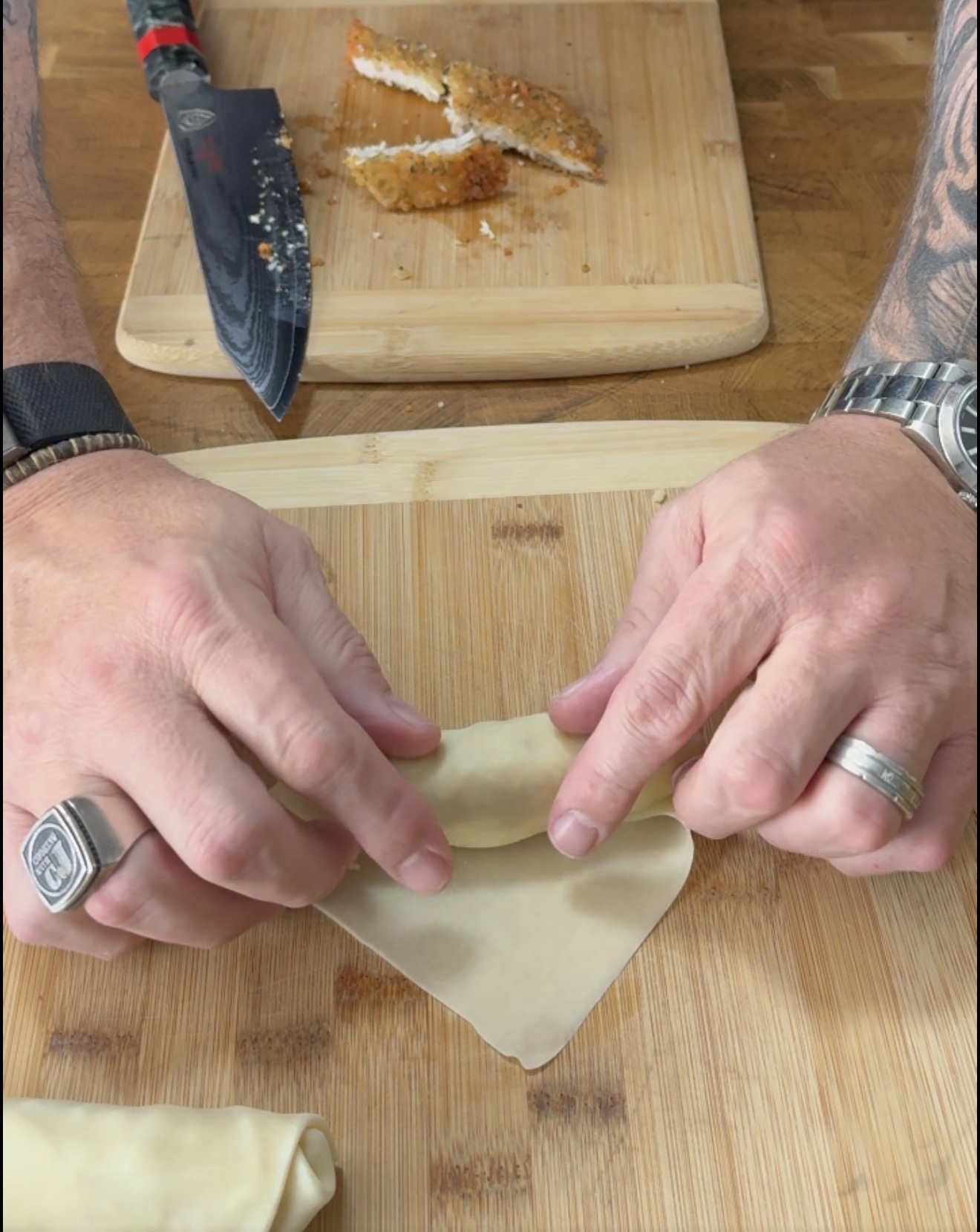 A persons hands roll a filling into a square wrapper on a wooden cutting board, with a knife and sliced breaded chicken nearby.