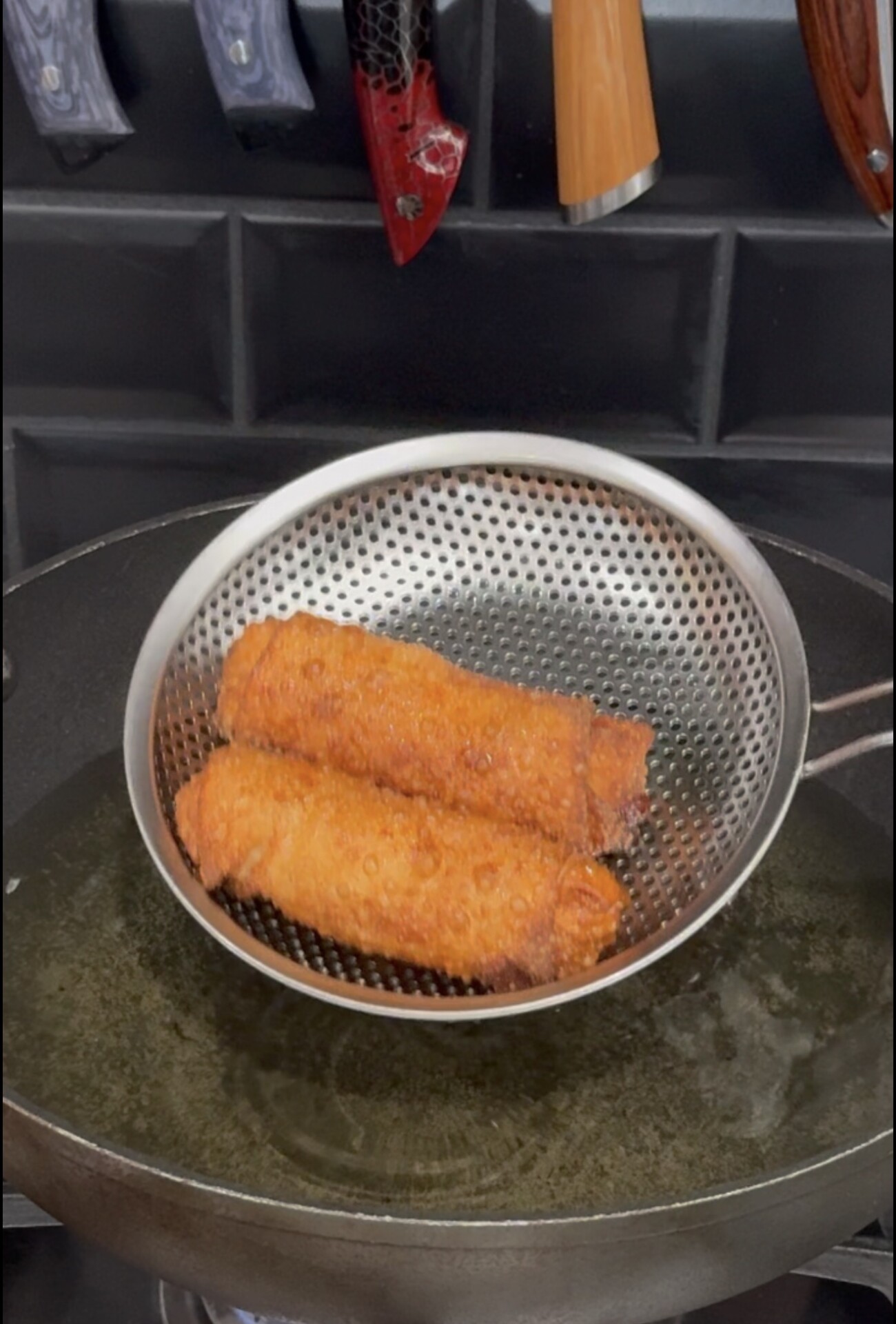Two fried egg rolls in a metal mesh strainer being lifted from a pan of hot oil.