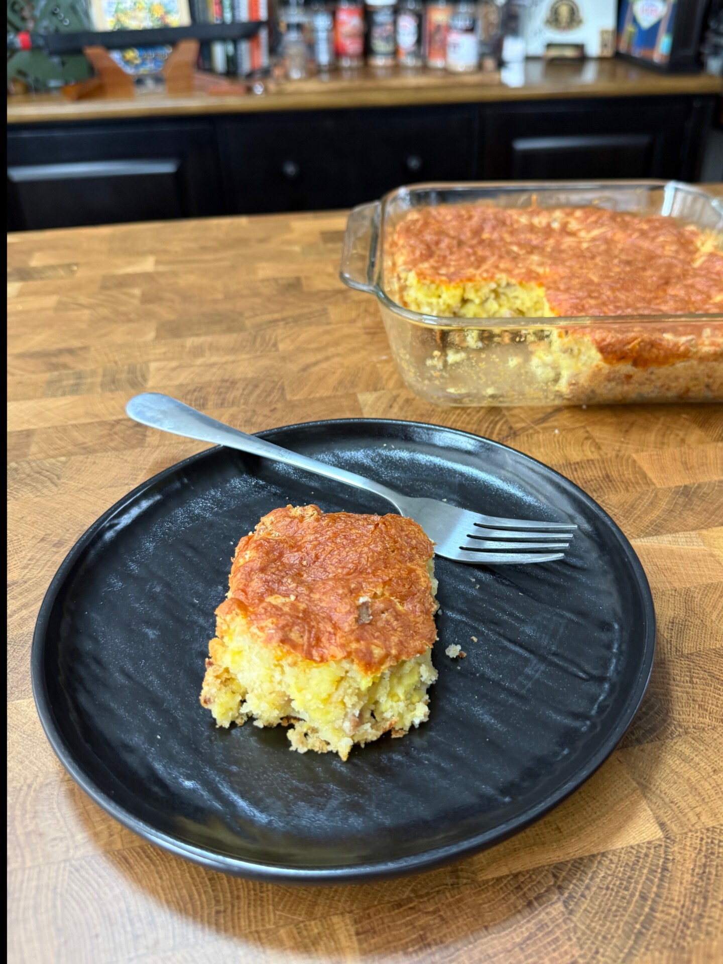 A slice of golden-brown cornbread on a black plate with a fork, next to a glass baking dish containing the rest of the cornbread.