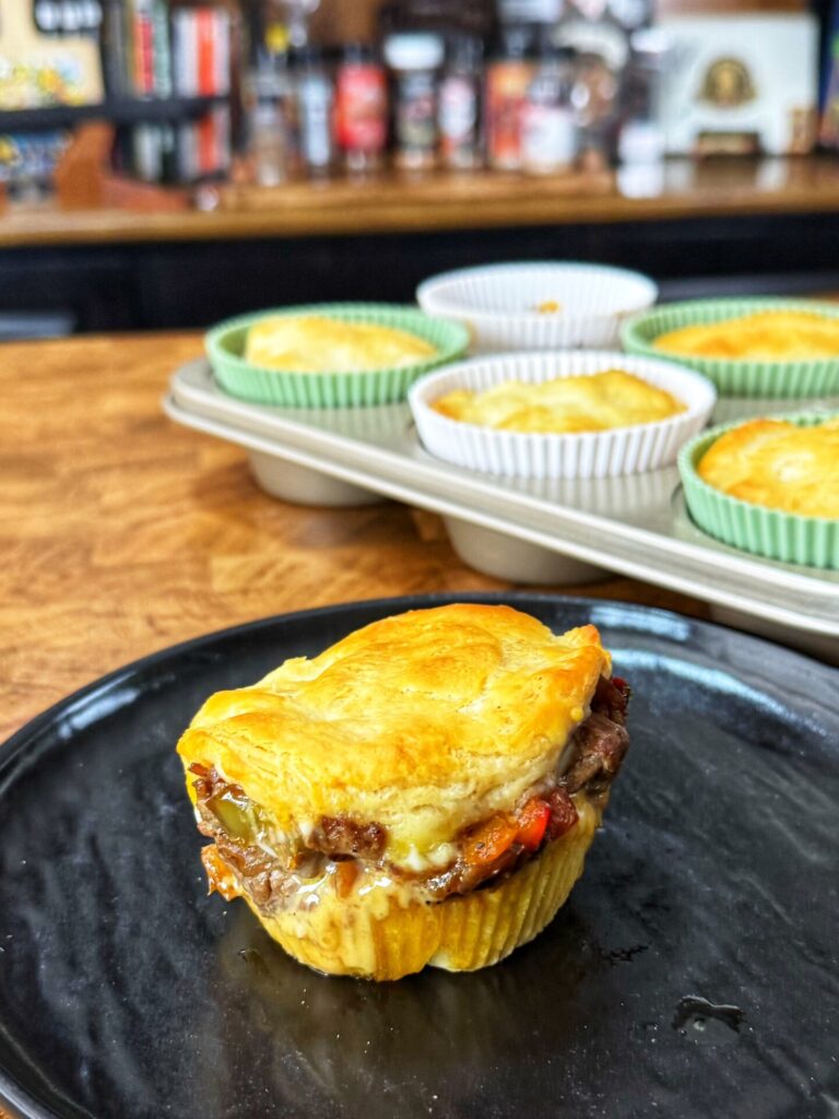 A single meat-filled pot pie in a muffin tin liner on a black plate, with more pies in a baking tray in the background.