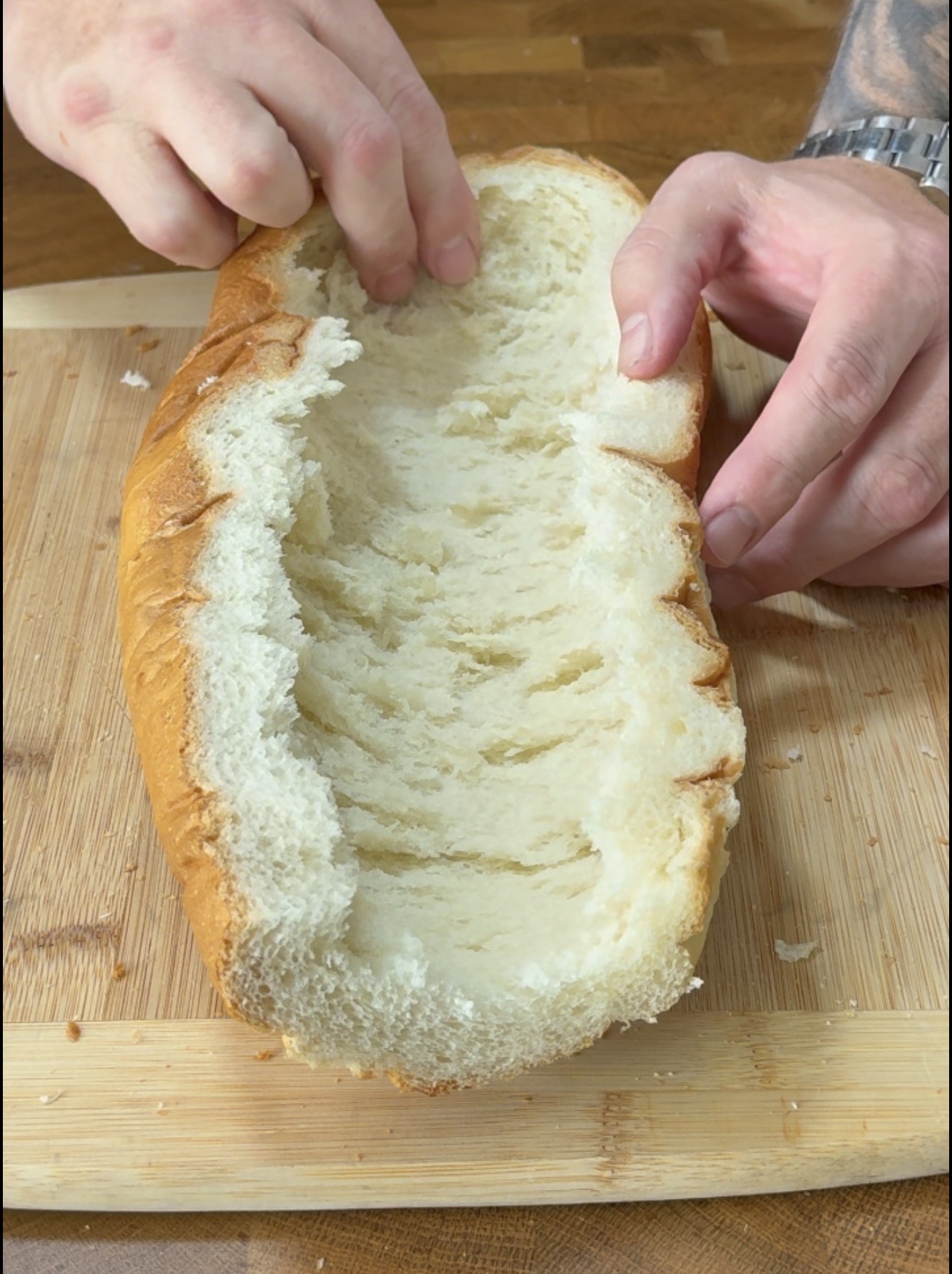 Hands pulling the soft interior out of a hollowed-out loaf of bread on a wooden cutting board.