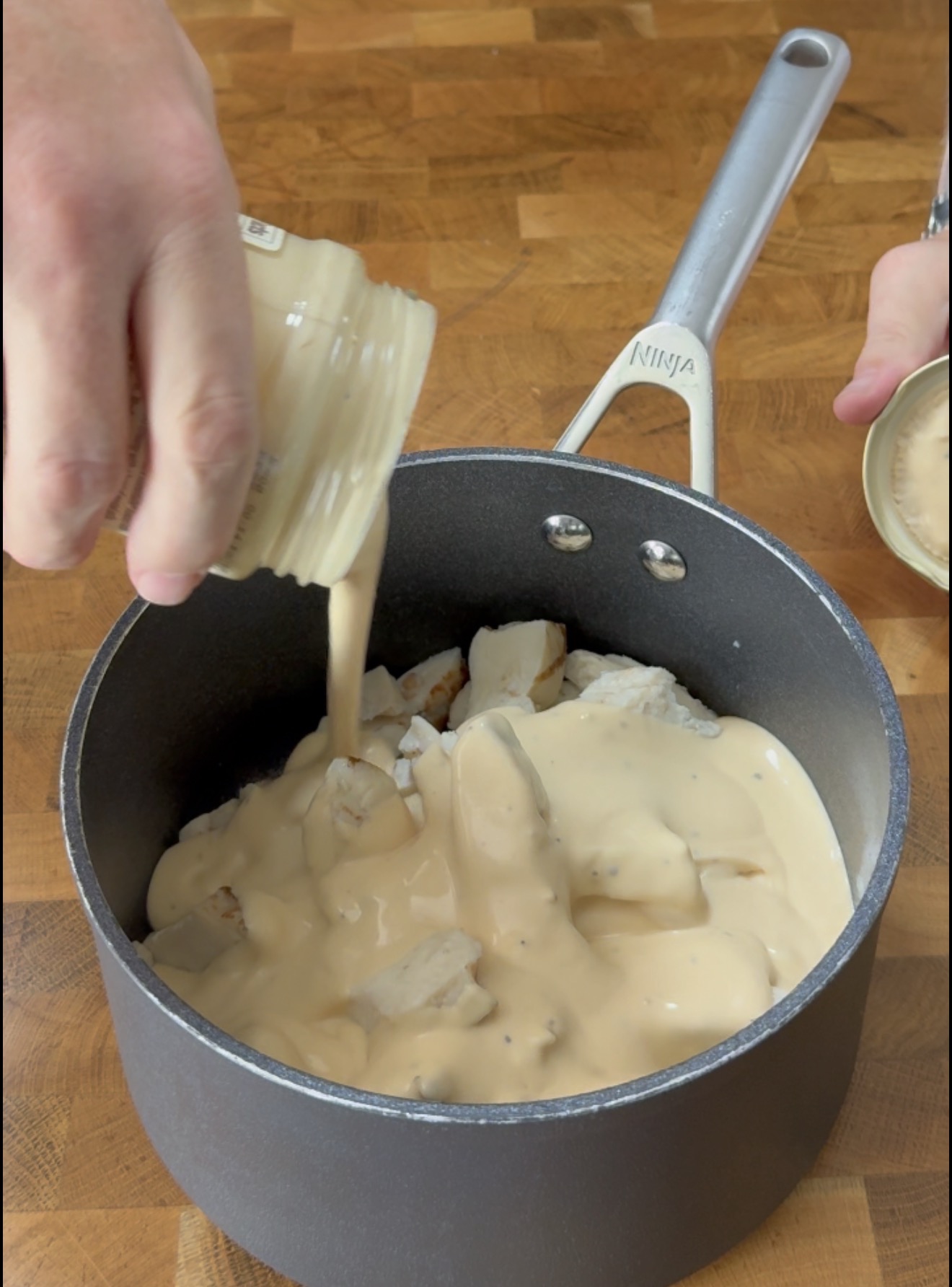 A hand pours a creamy sauce from a jar into a pot containing chopped chicken.
