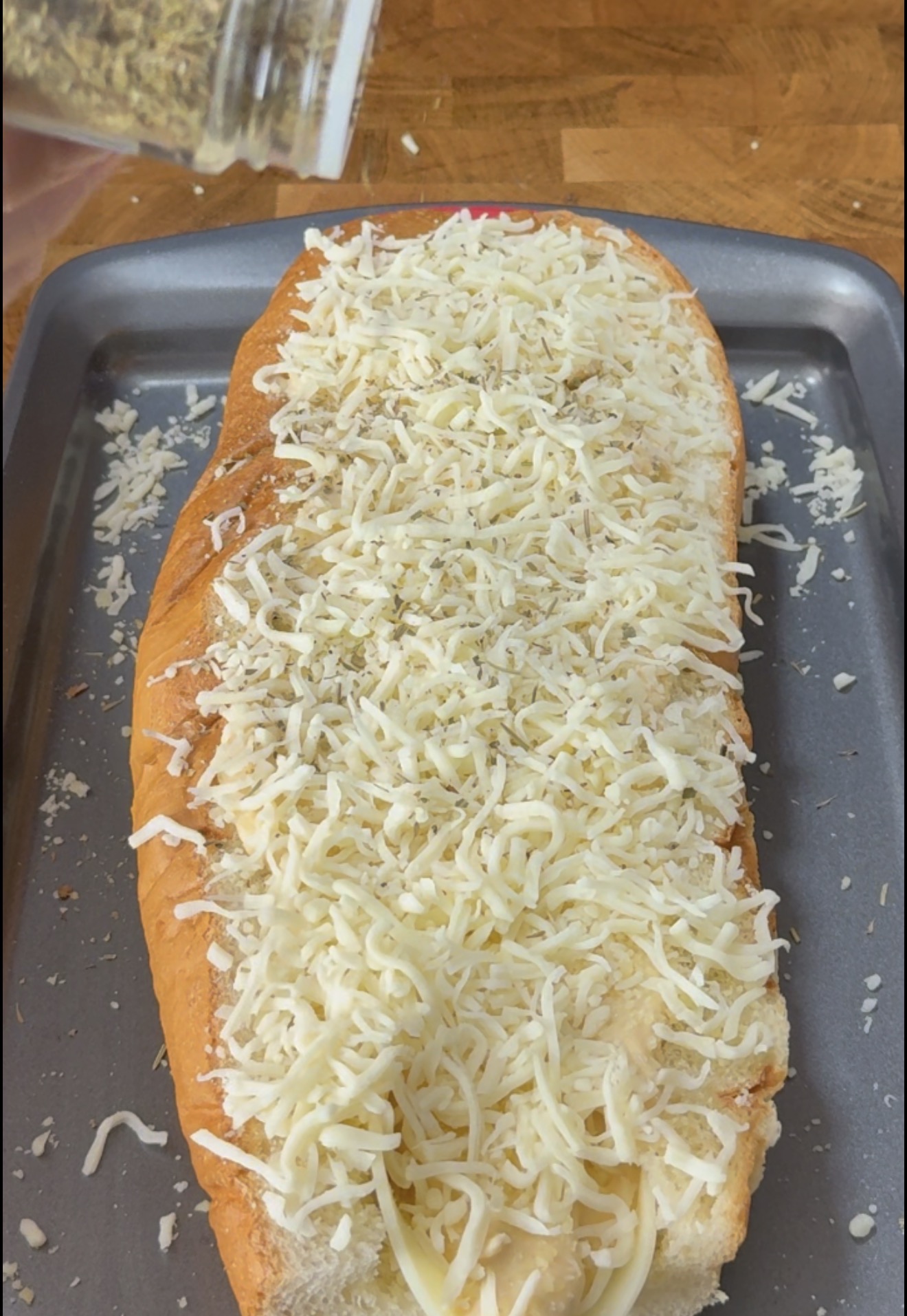 A hand shakes dried herbs onto a loaf of bread topped with shredded cheese on a baking sheet.